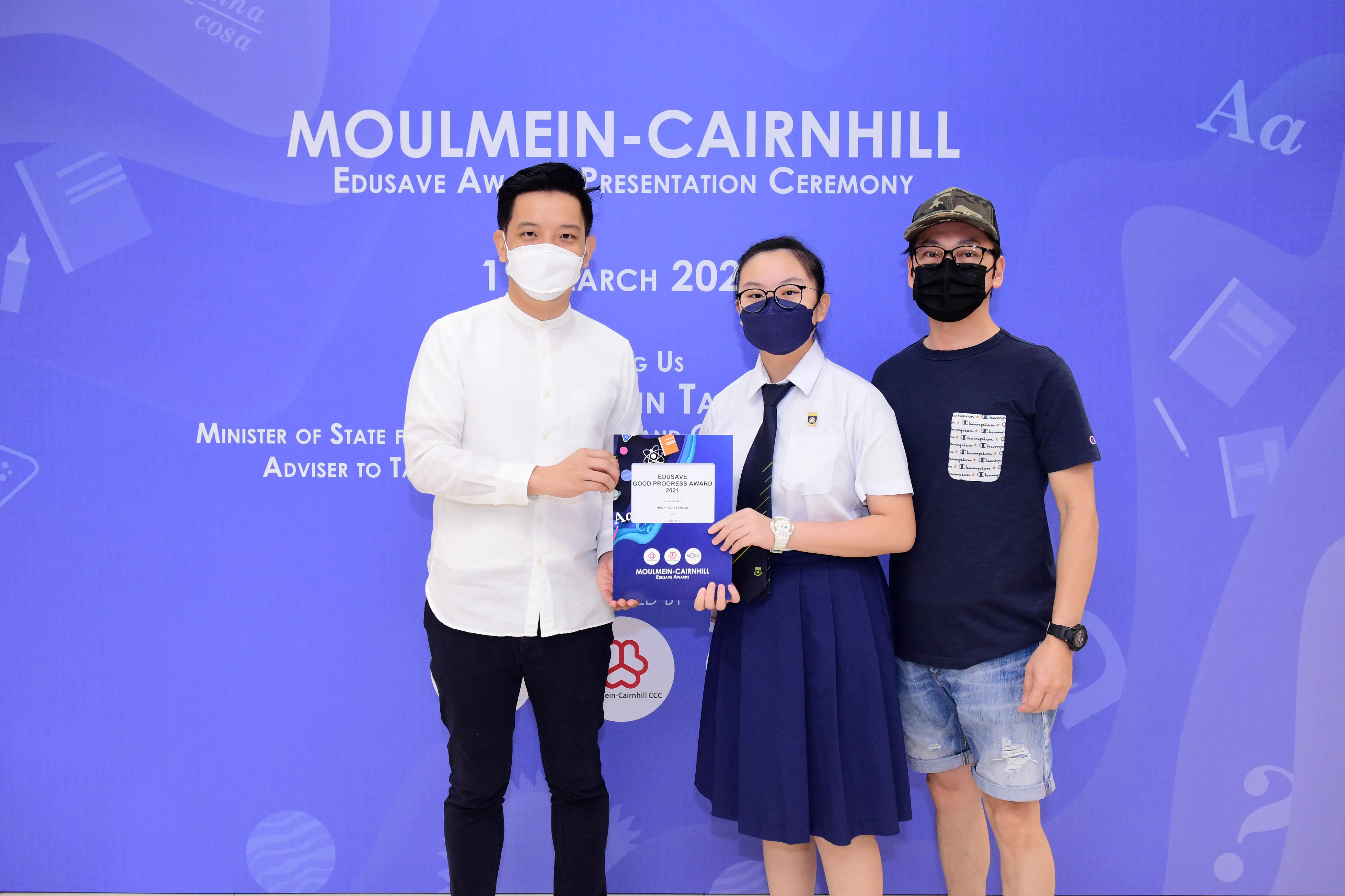 Three masked people pose at the Moulmein-Cairnhill Edusave Award Presentation Ceremony, with a student holding an award.