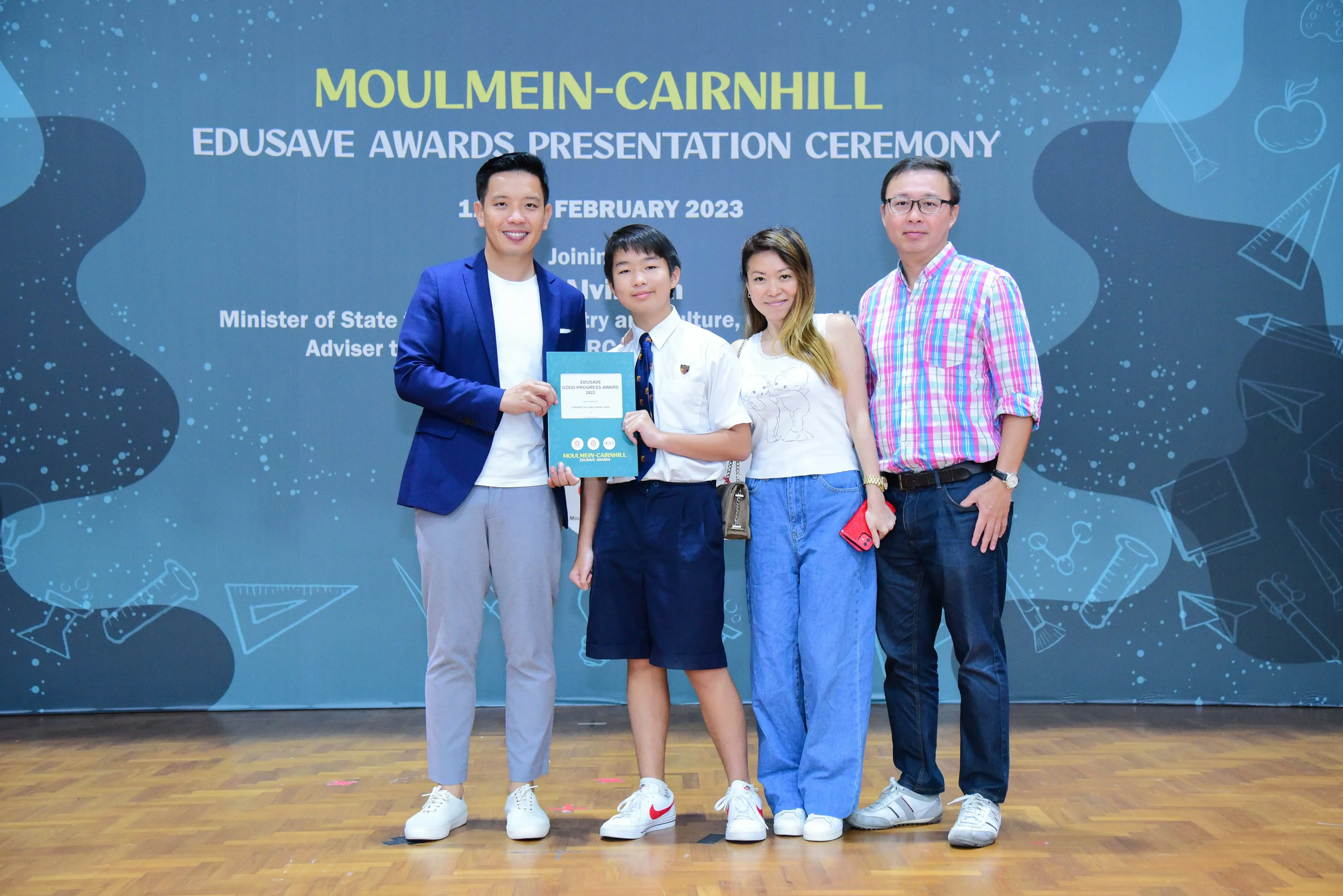 Four people pose at the Moulmein-Cairnhill Edusave Awards Presentation Ceremony, a student holding a teal award folder.