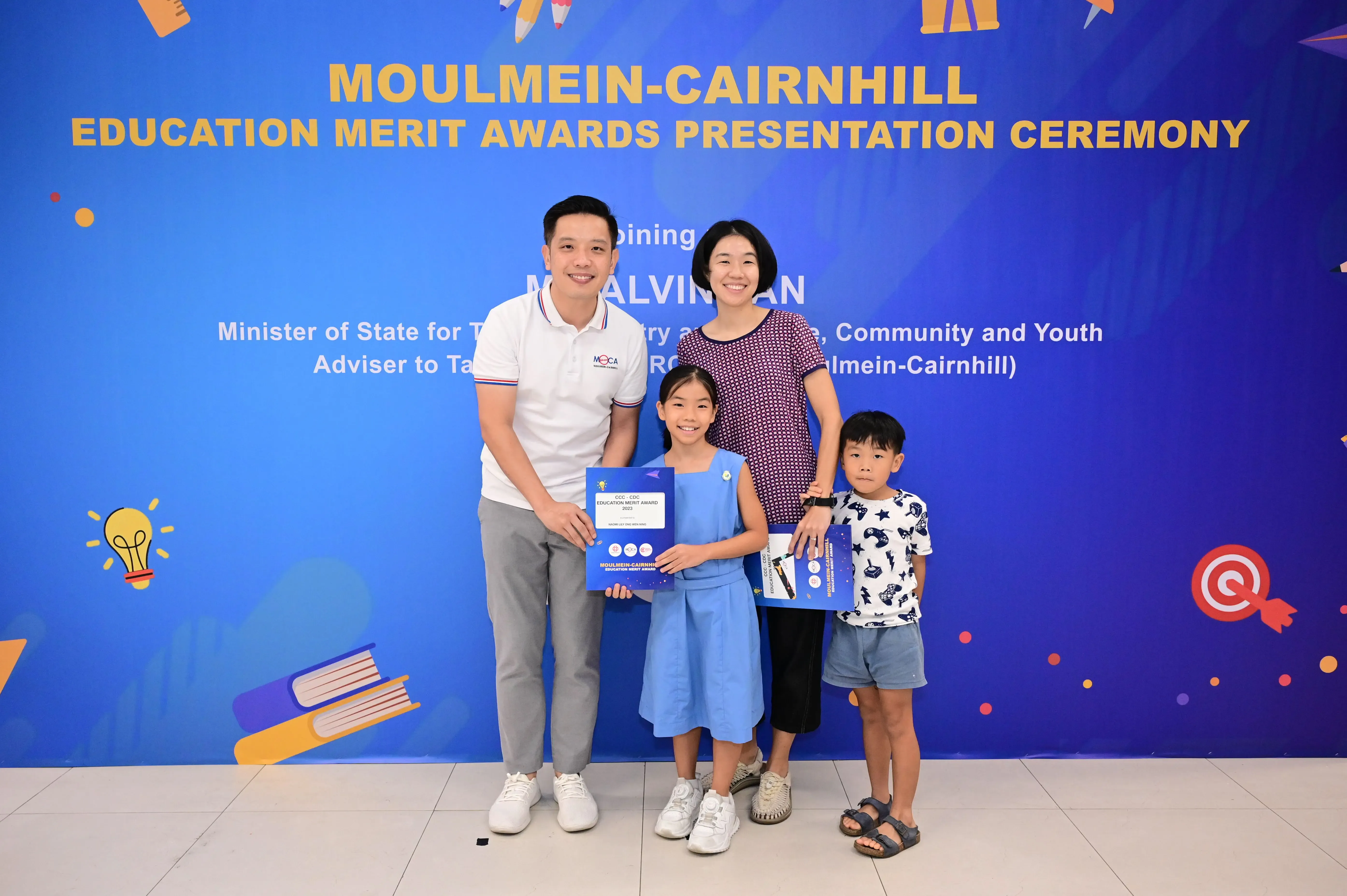 Family of four poses before a Moulmein-Cairnhill Education Merit Awards banner, girl holding a blue award certificate.
