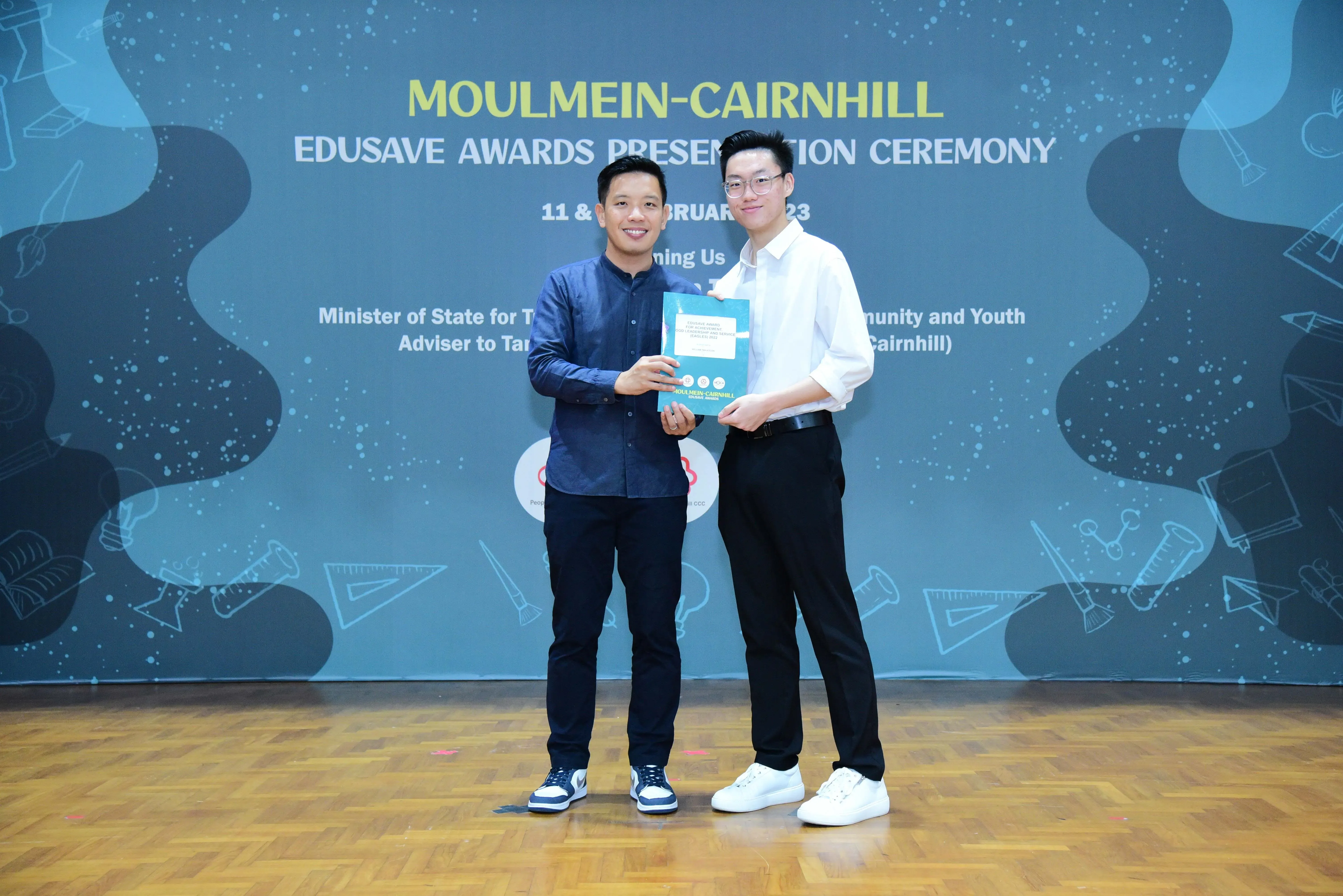 Two men hold a certificate together at the Moulmein-Cairnhill Edusave Awards Presentation Ceremony backdrop.