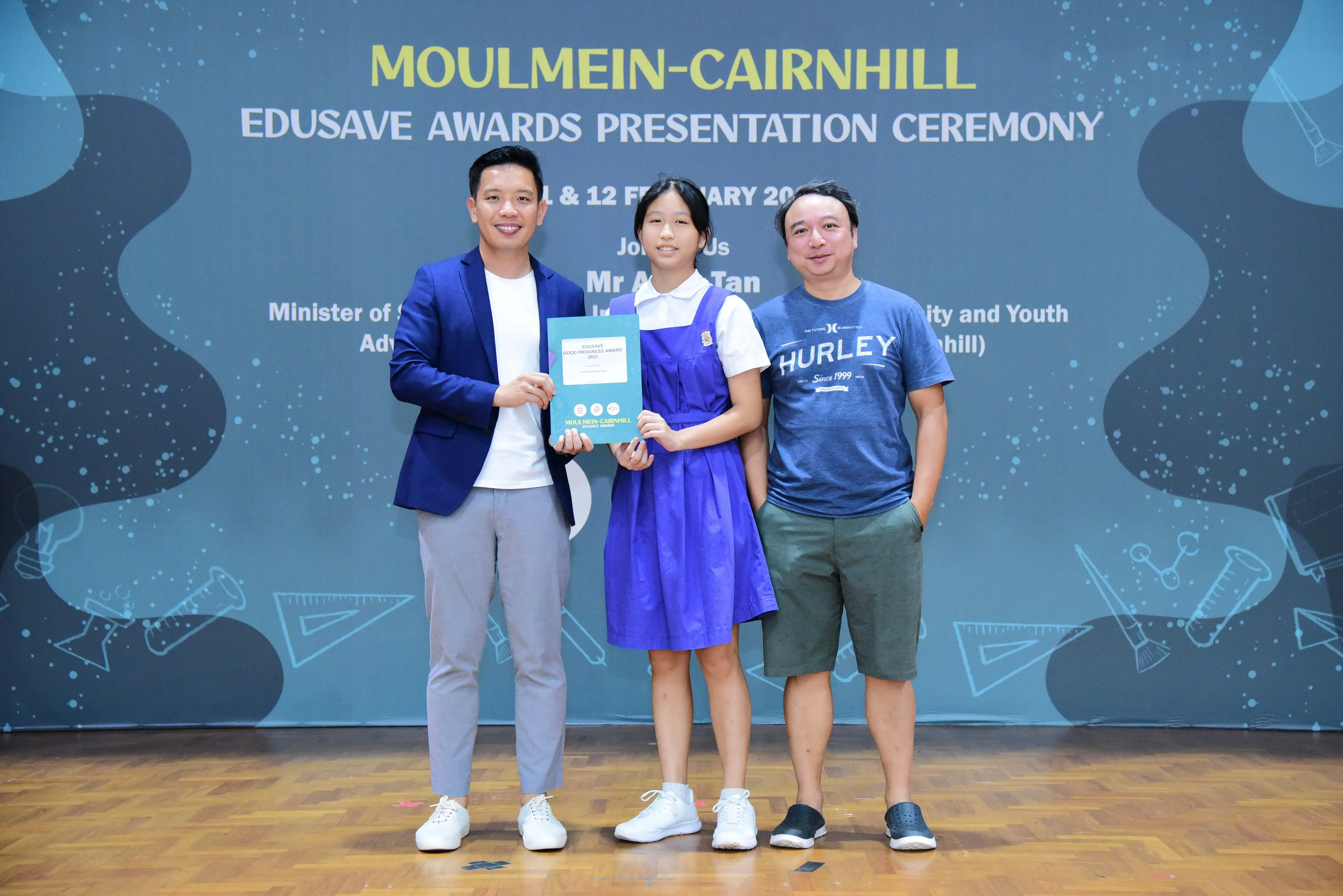 Three people pose at the Moulmein-Cairnhill Edusave Awards Presentation Ceremony, with a girl in uniform holding an award.