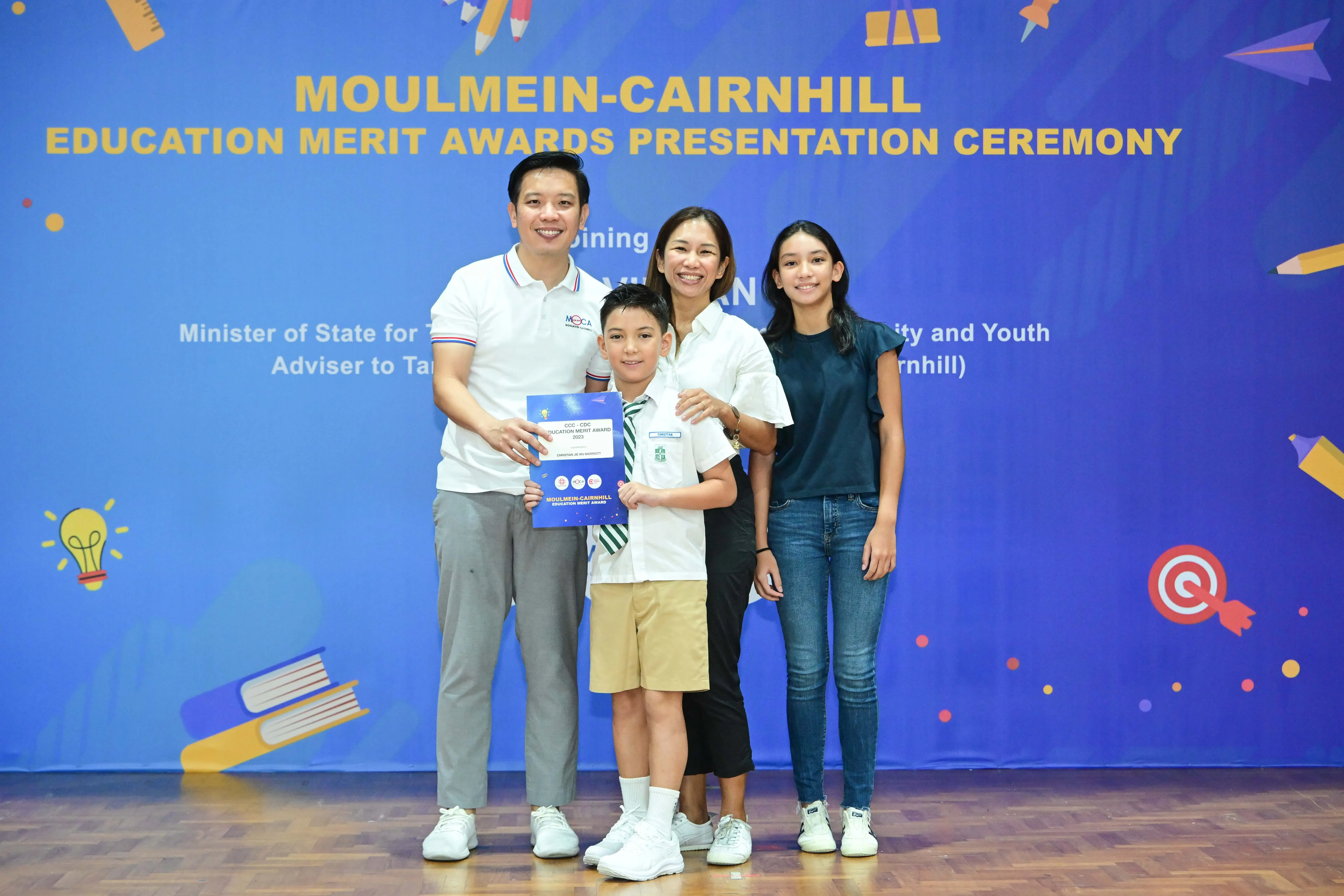 Family of four poses before a Moulmein-Cairnhill Education Merit Awards banner; a boy holds a blue certificate.