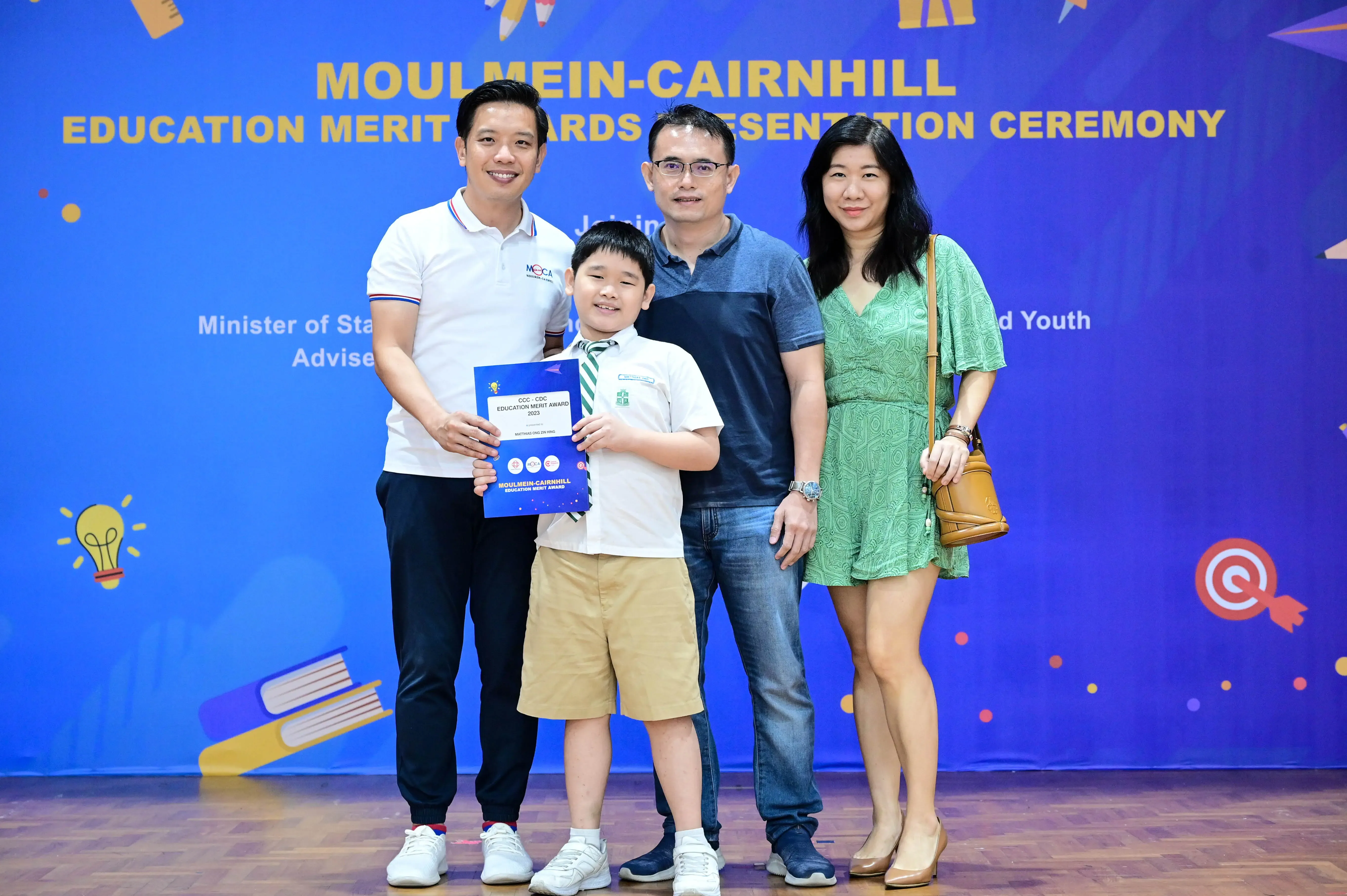 A young boy holds an award certificate with three adults at the Moulmein-Cairnhill Education Merit Awards Ceremony.