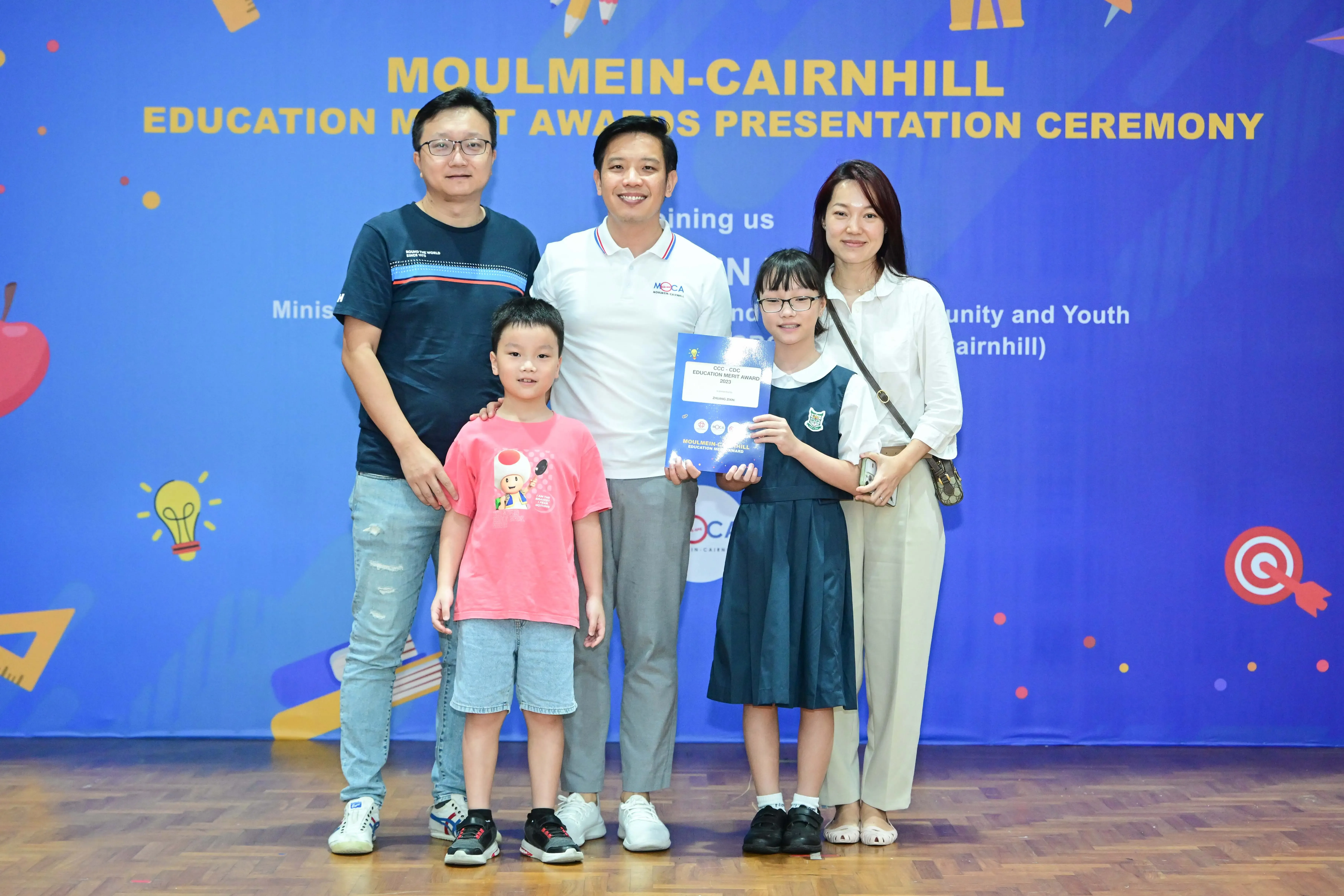 Five people pose in front of a Moulmein-Cairnhill Education Merit Awards Presentation Ceremony banner.