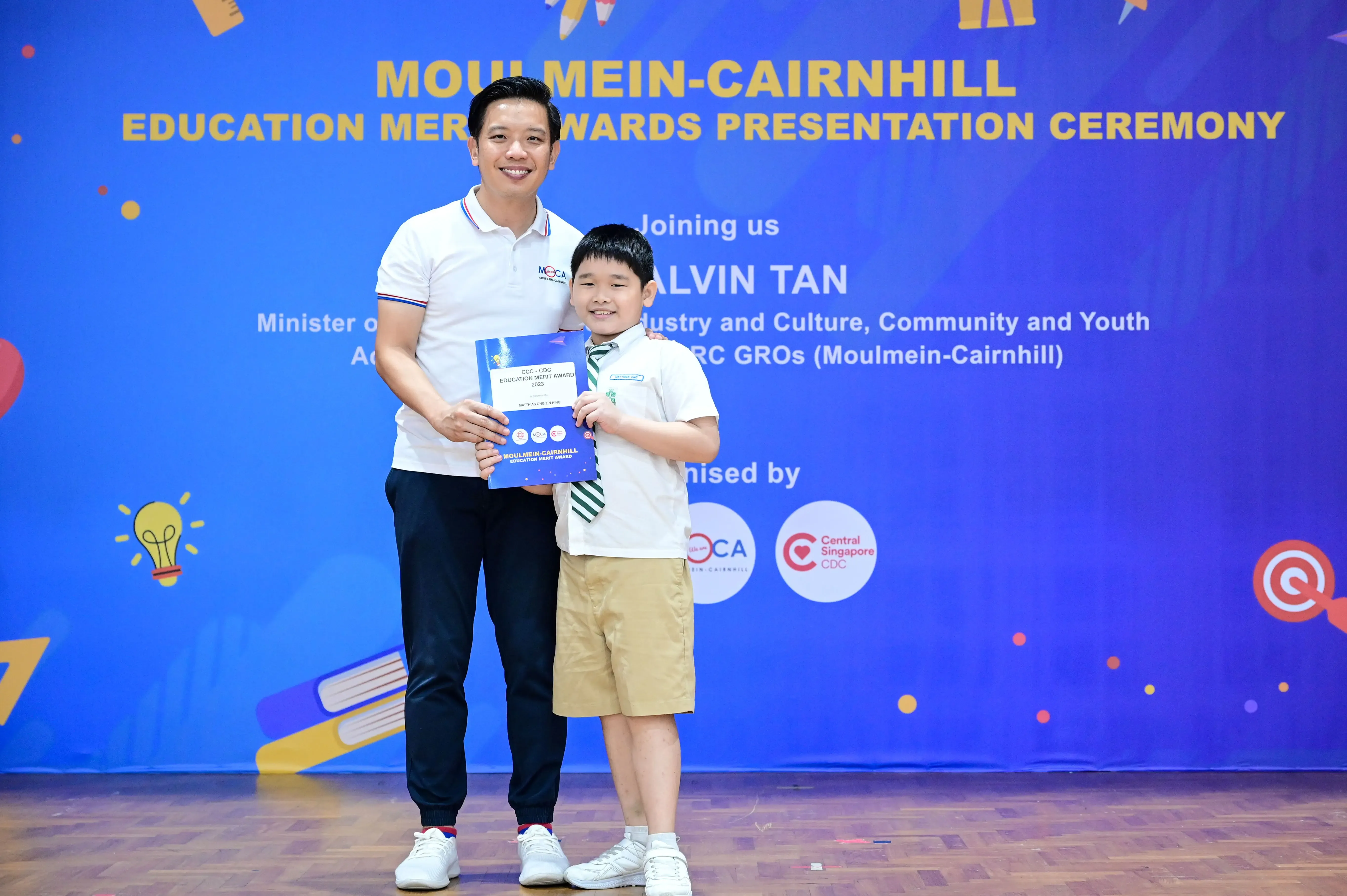 An adult man and a young boy hold an award certificate at the Moulmein-Cairnhill Education Merit Awards ceremony.