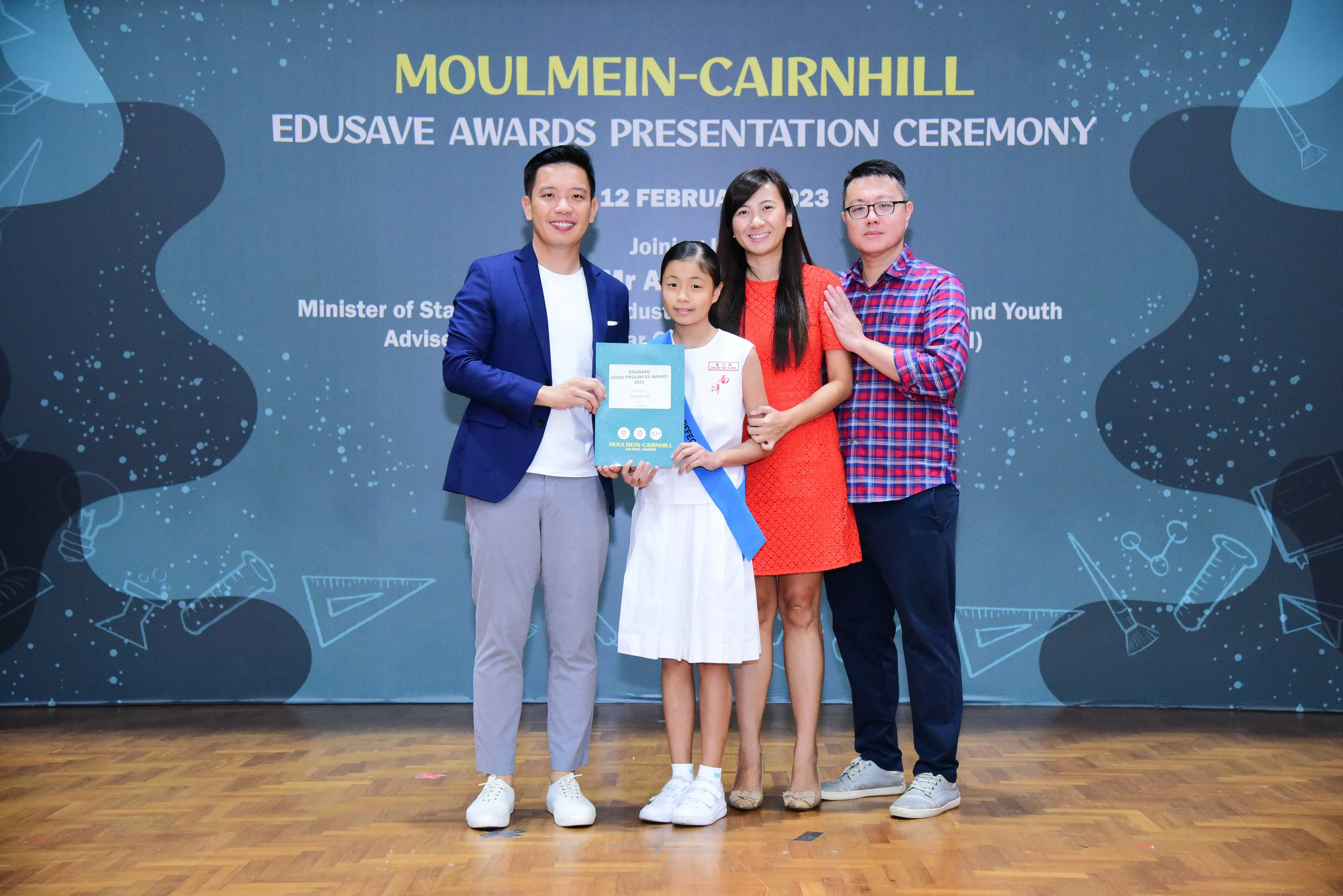 Four people pose at the Moulmein-Cairnhill Edusave Awards Presentation Ceremony, with a girl holding an award certificate.