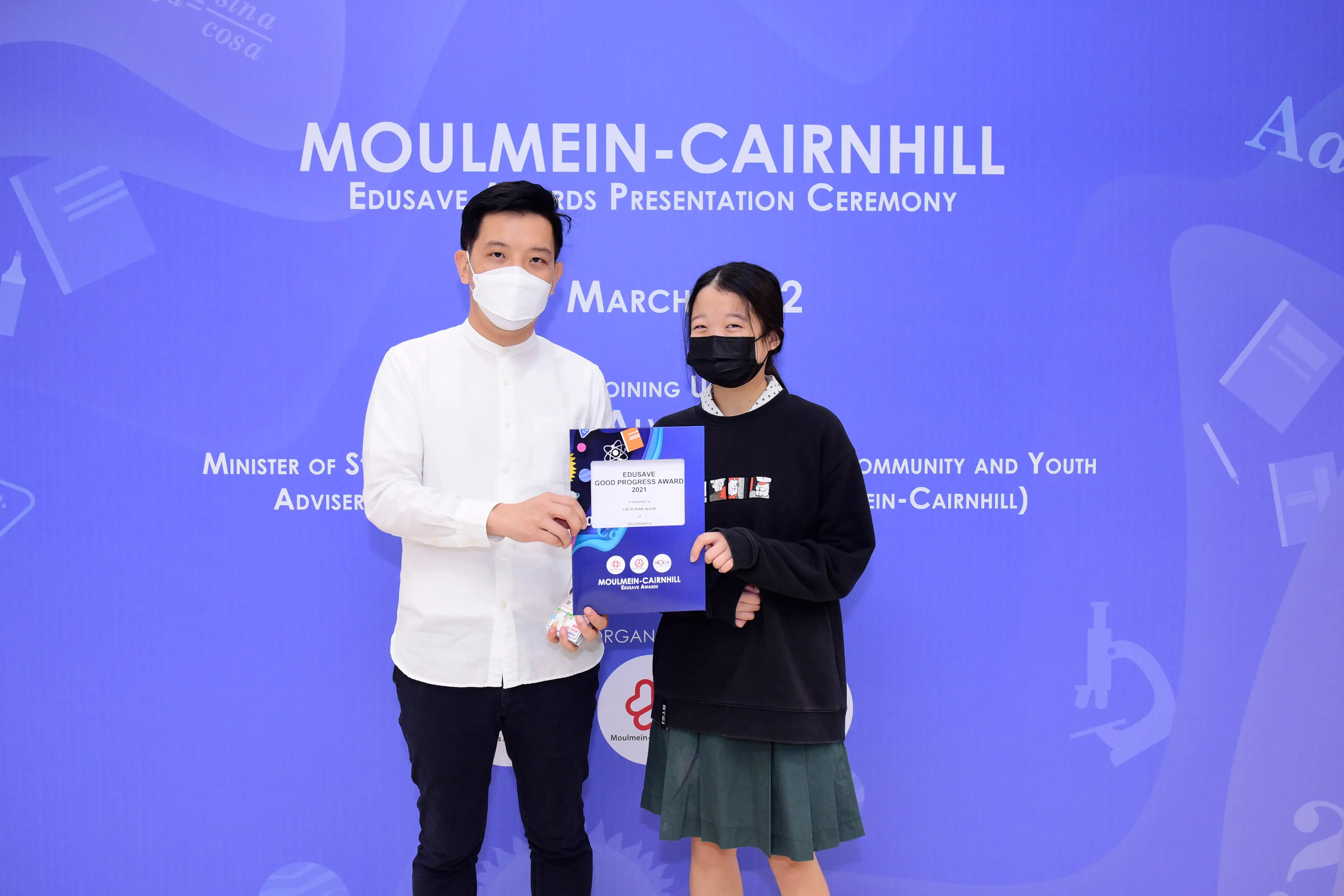 Two masked people stand before a blue Moulmein-Cairnhill Edusave Awards Presentation Ceremony banner, holding a certificate.