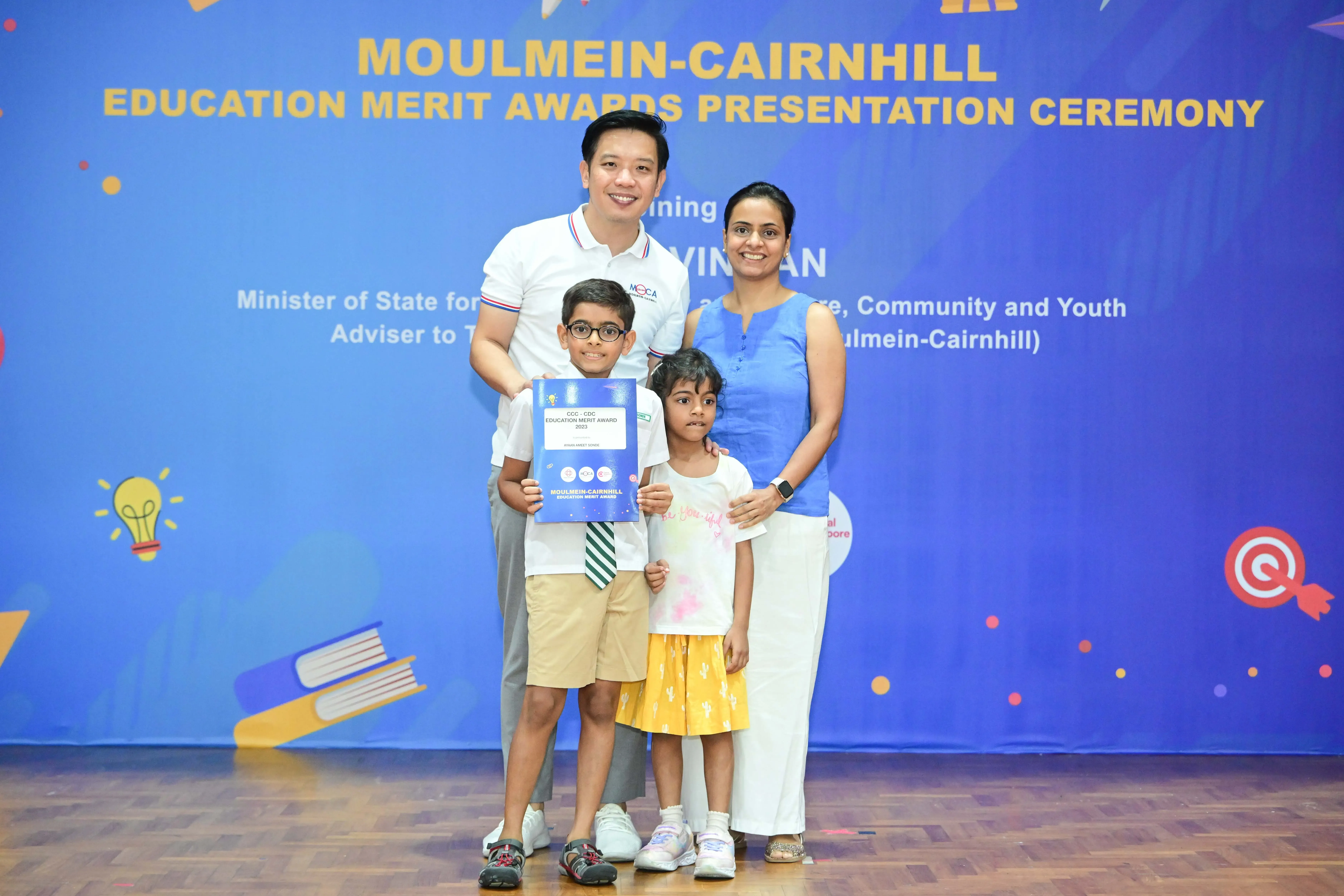 Family of four posing with an Education Merit Award certificate at the Moulmein-Cairnhill ceremony backdrop.