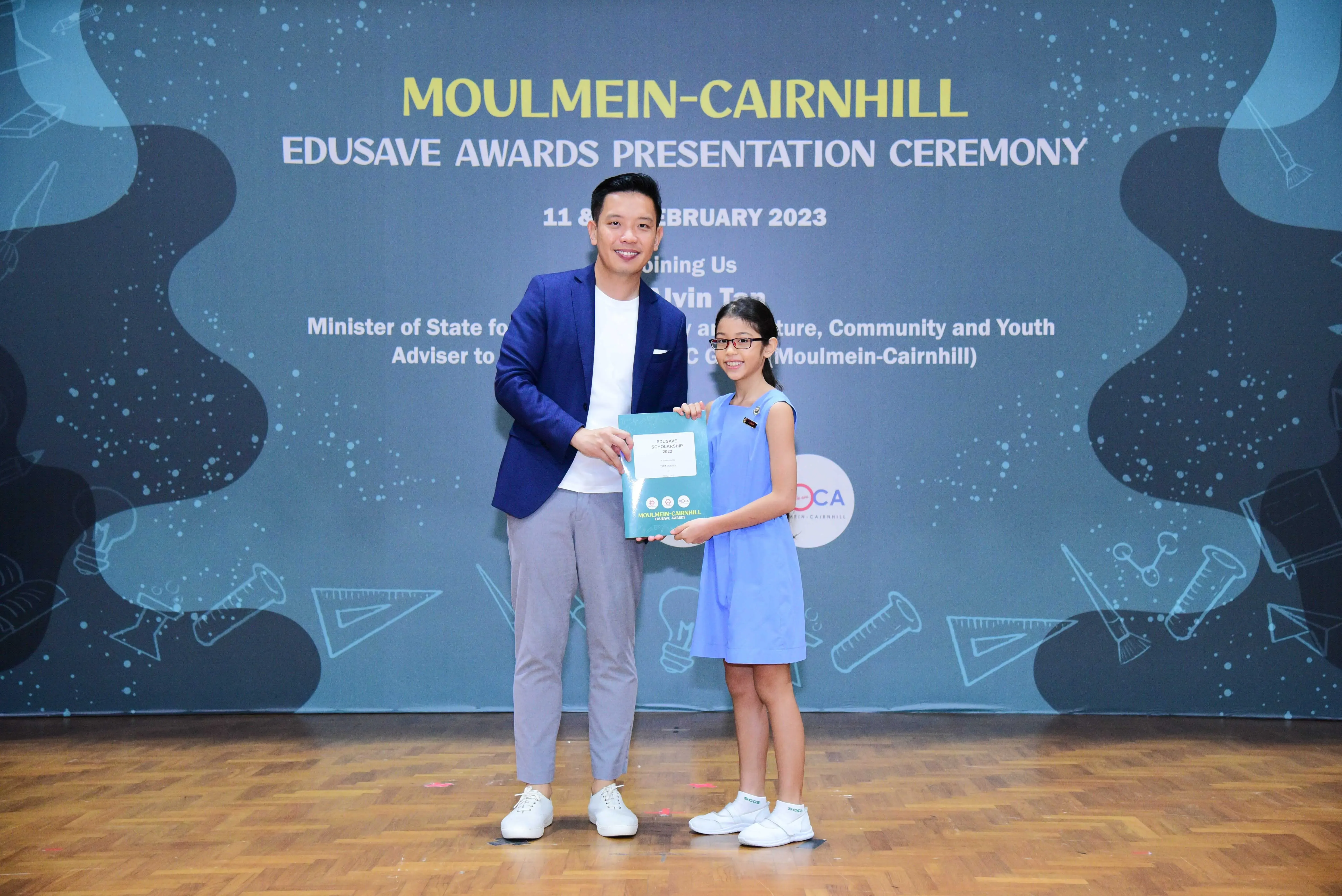 A man in a blue blazer and a young girl in a blue dress hold an award folder at the Moulmein-Cairnhill Edusave Awards 2023.