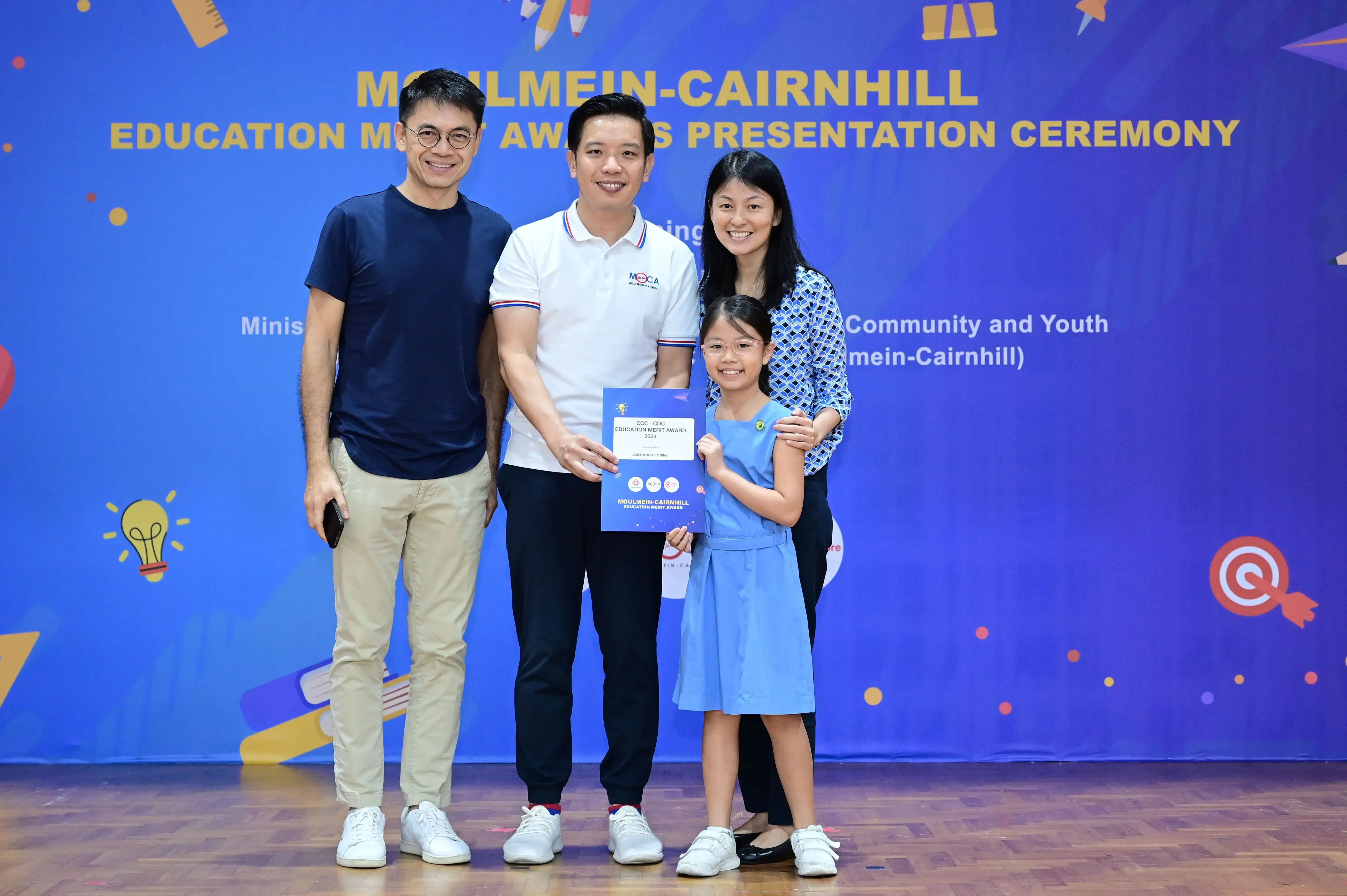 Three adults and a young girl pose holding a certificate at the Moulmein-Cairnhill Education Merit Awards ceremony.