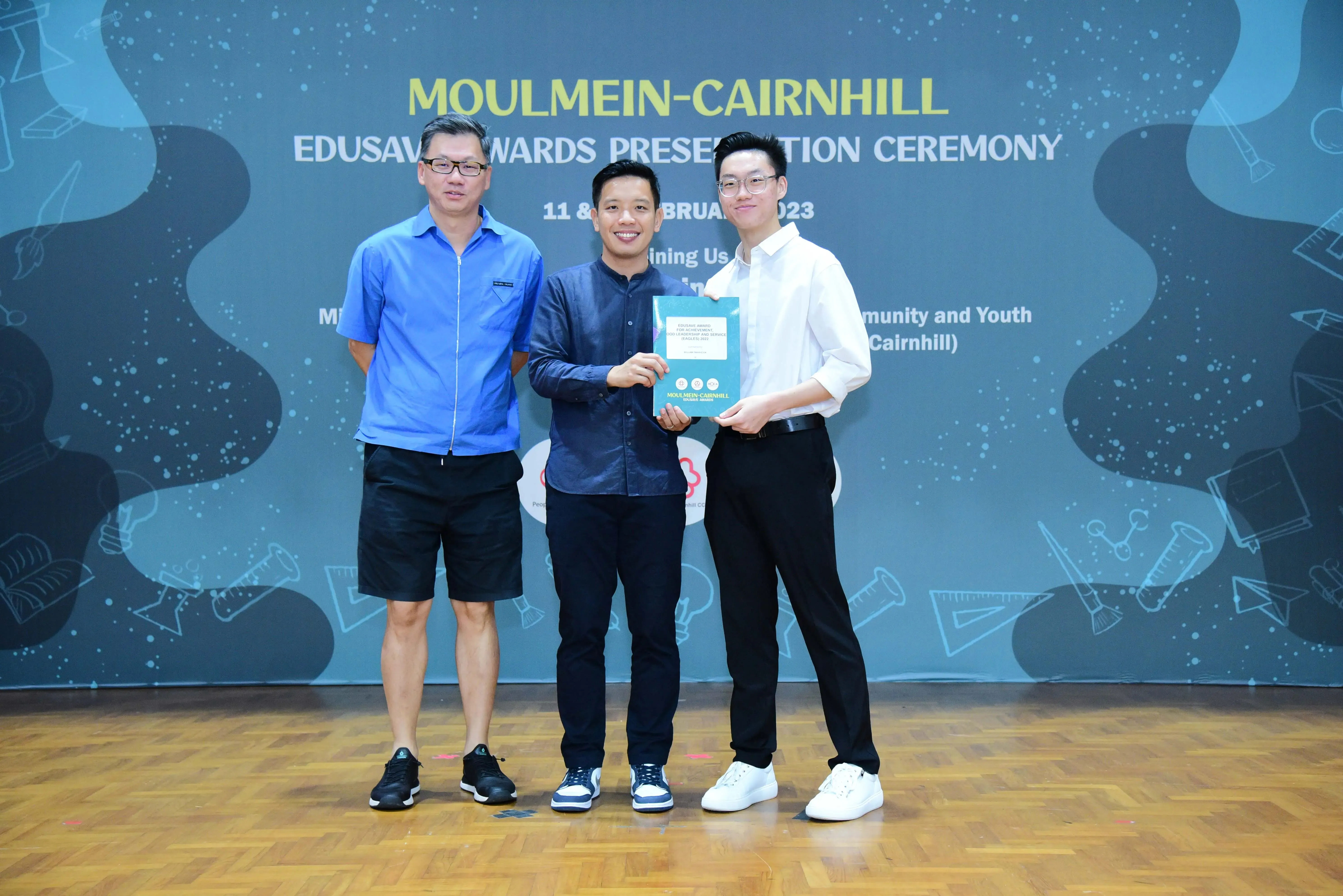 Three men pose together holding a booklet at the Moulmein-Cairnhill Edusave Awards Presentation Ceremony 2023.