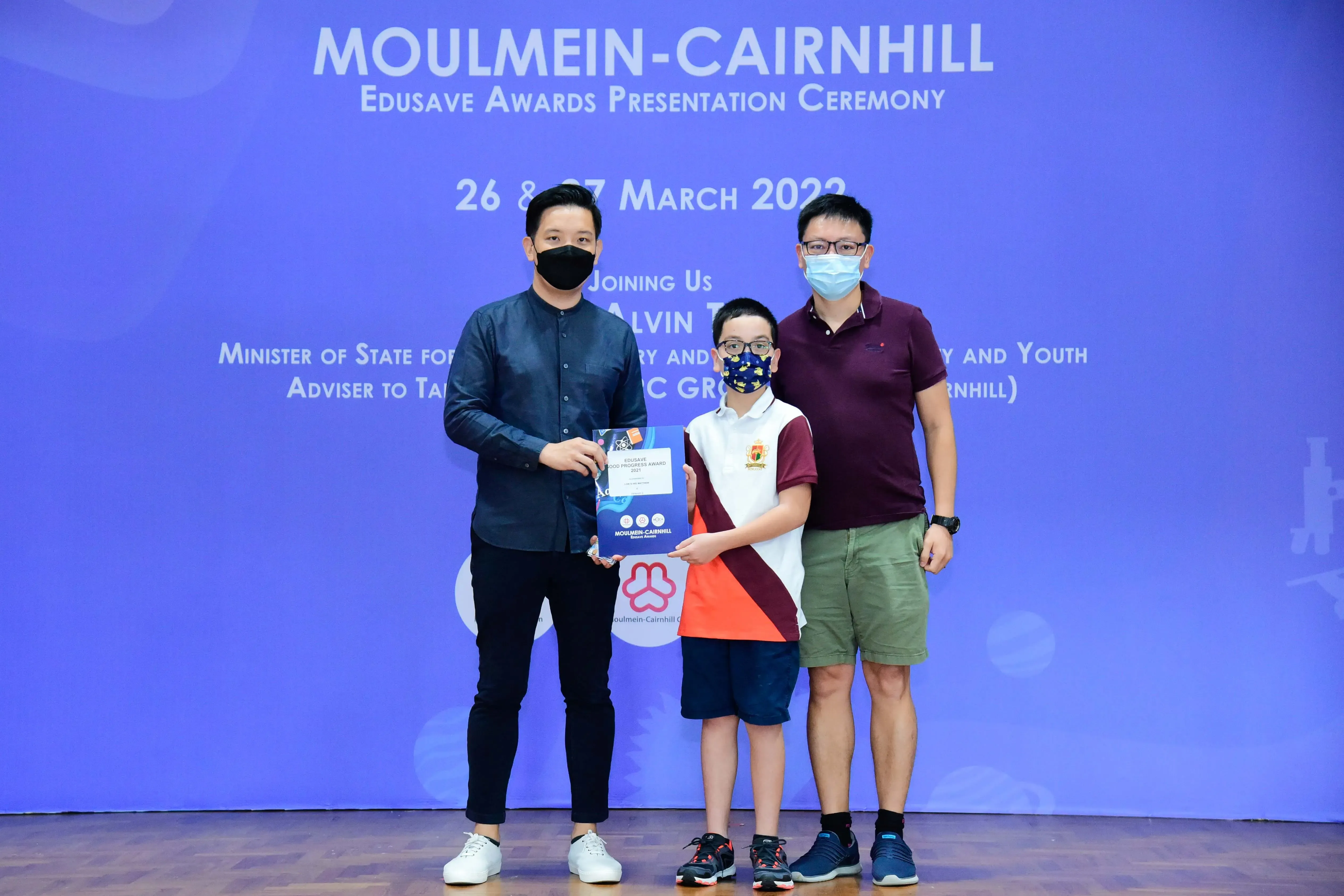 Two masked adults and a masked child hold an award at the Moulmein-Cairnhill Edusave Awards Ceremony, March 2022.
