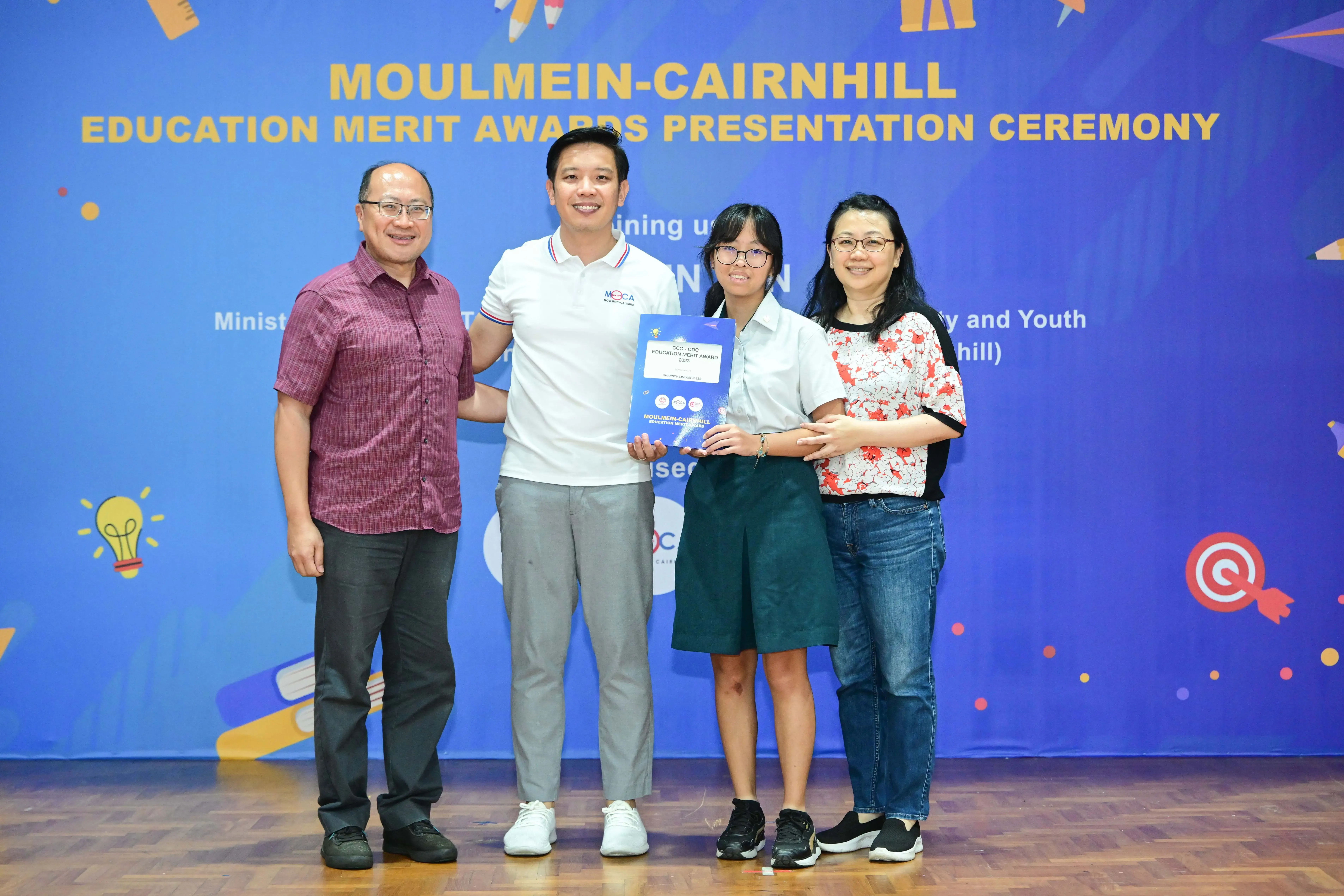 Four people pose smiling at the Moulmein-Cairnhill Education Merit Awards Presentation Ceremony, holding an award certificate.
