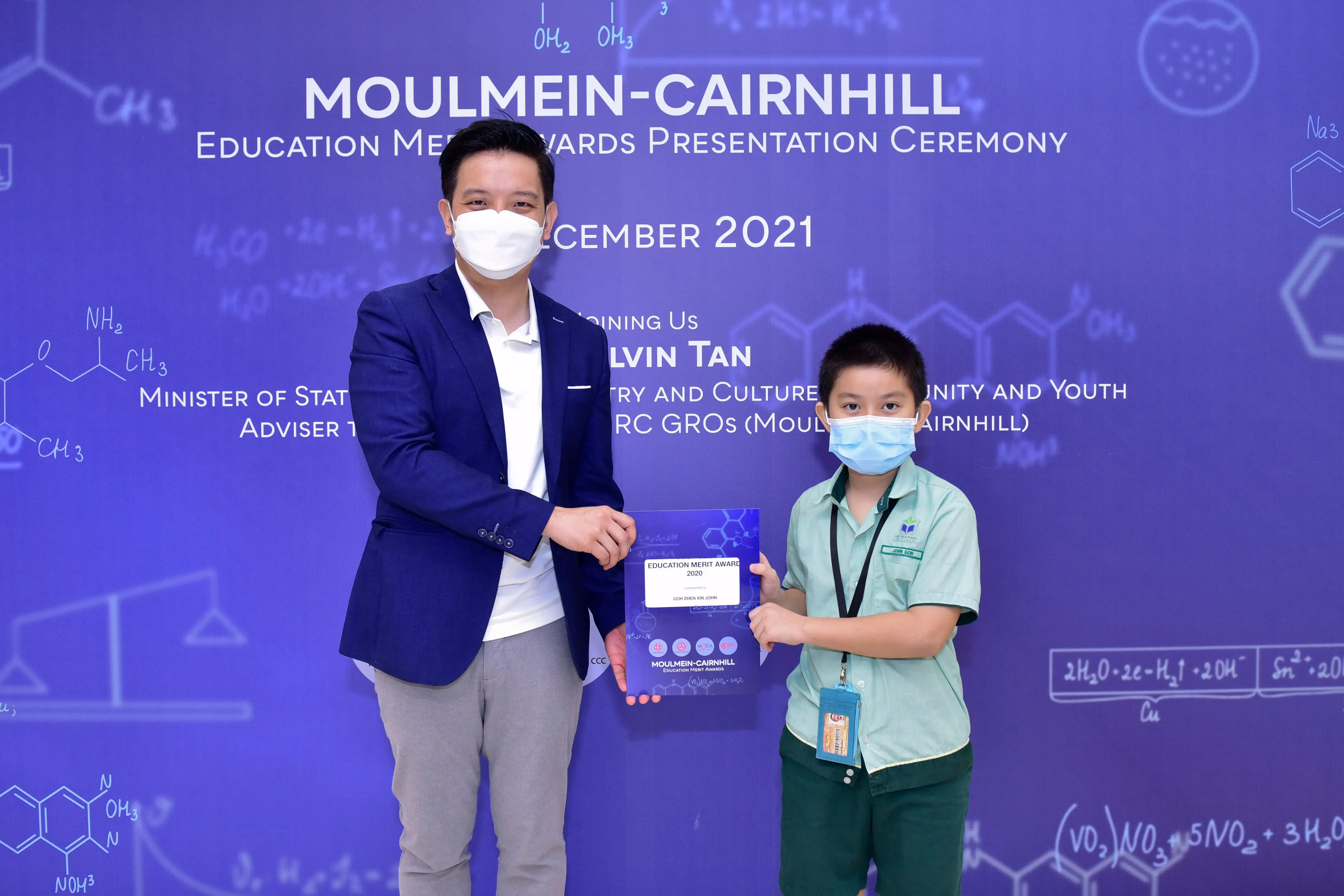 An adult and a young boy wearing masks hold an Education Merit Award certificate at the Moulmein-Cairnhill ceremony, December 2021.