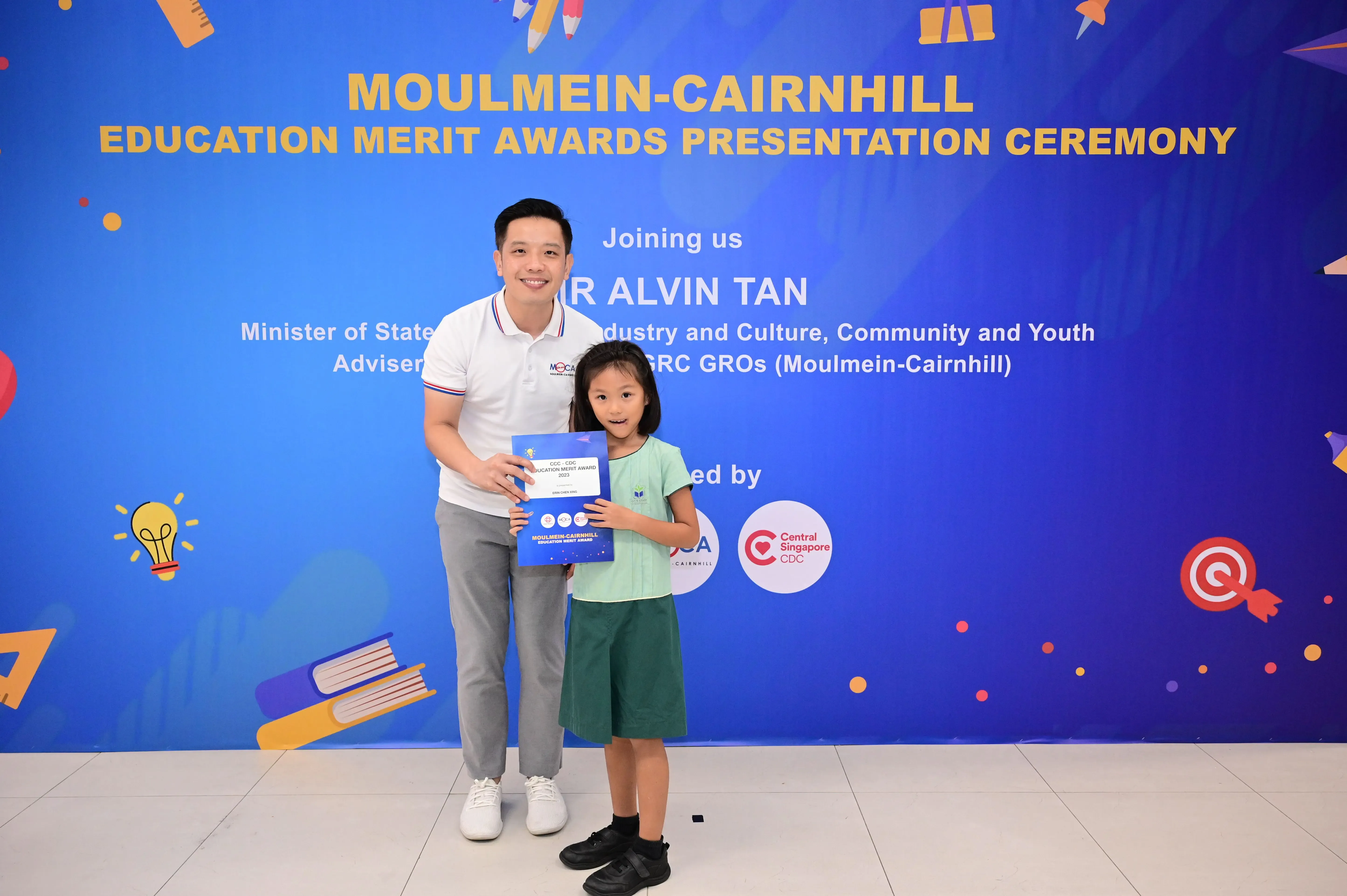 A man and a young girl hold a certificate in front of a Moulmein-Cairnhill Education Merit Awards banner.