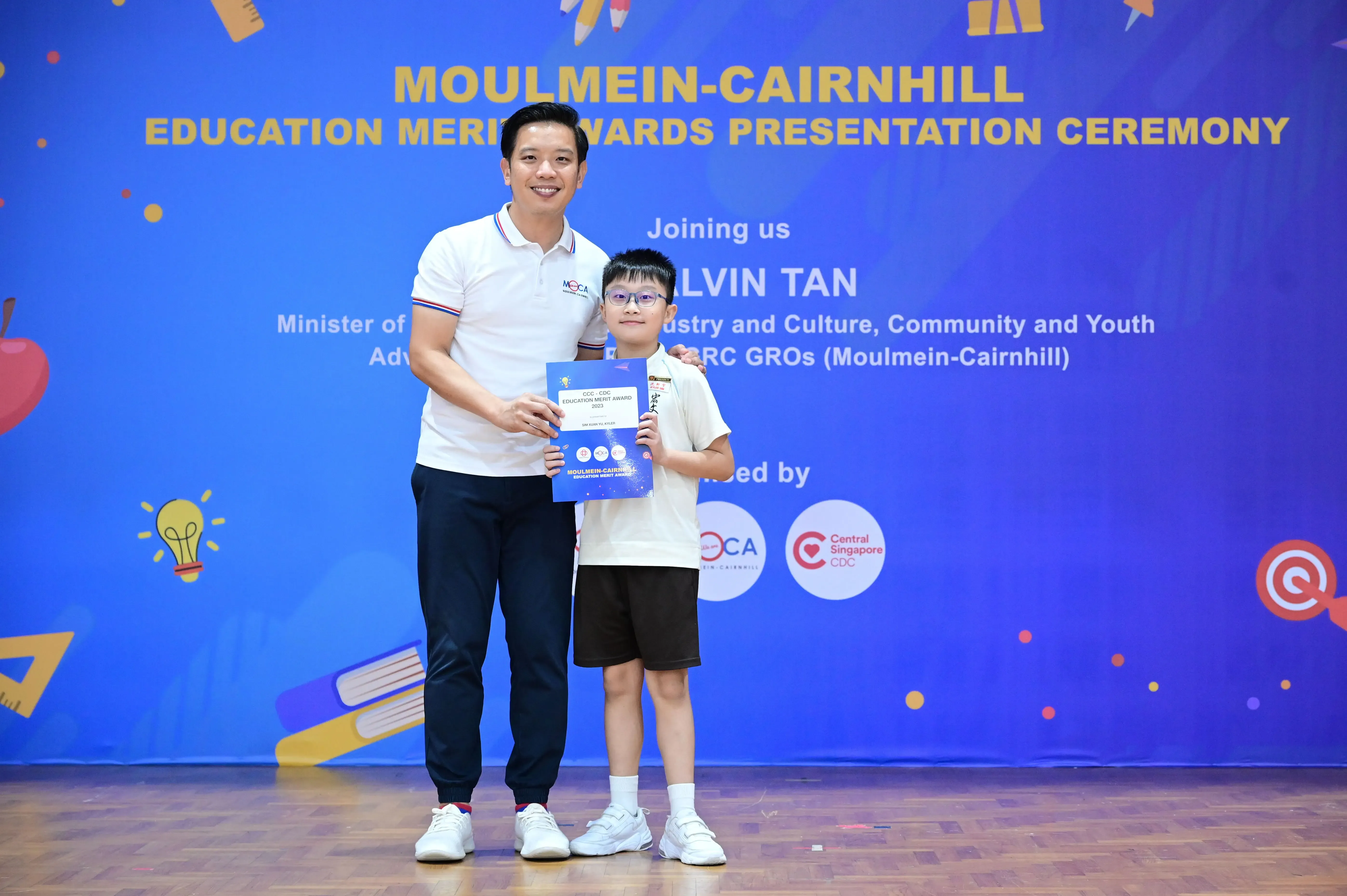 An adult man and a young boy hold a certificate together in front of a Moulmein-Cairnhill Education Merit Awards banner.