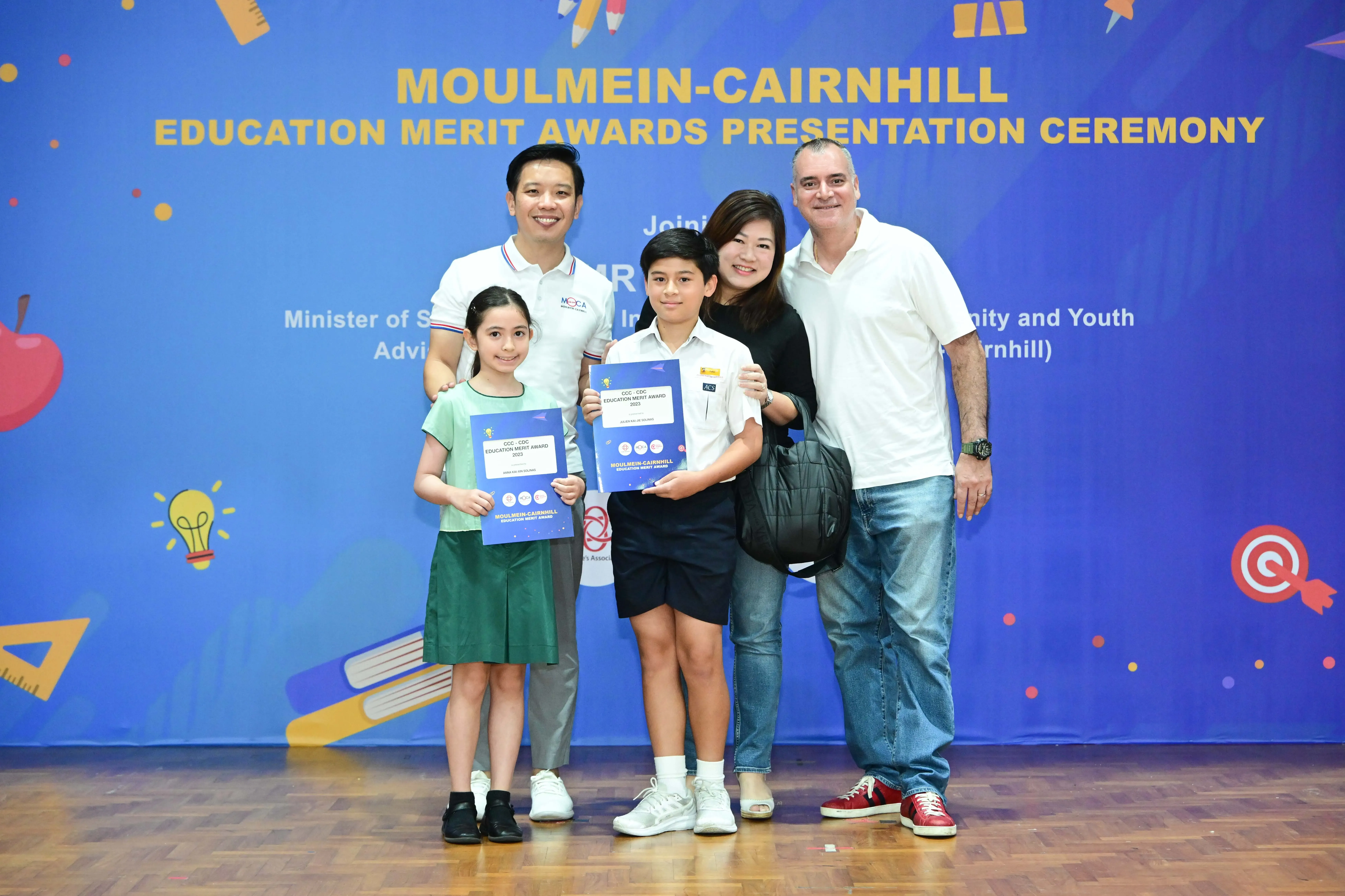 Two children holding blue merit award certificates pose with three adults at Moulmein-Cairnhill Education Merit Awards ceremony.