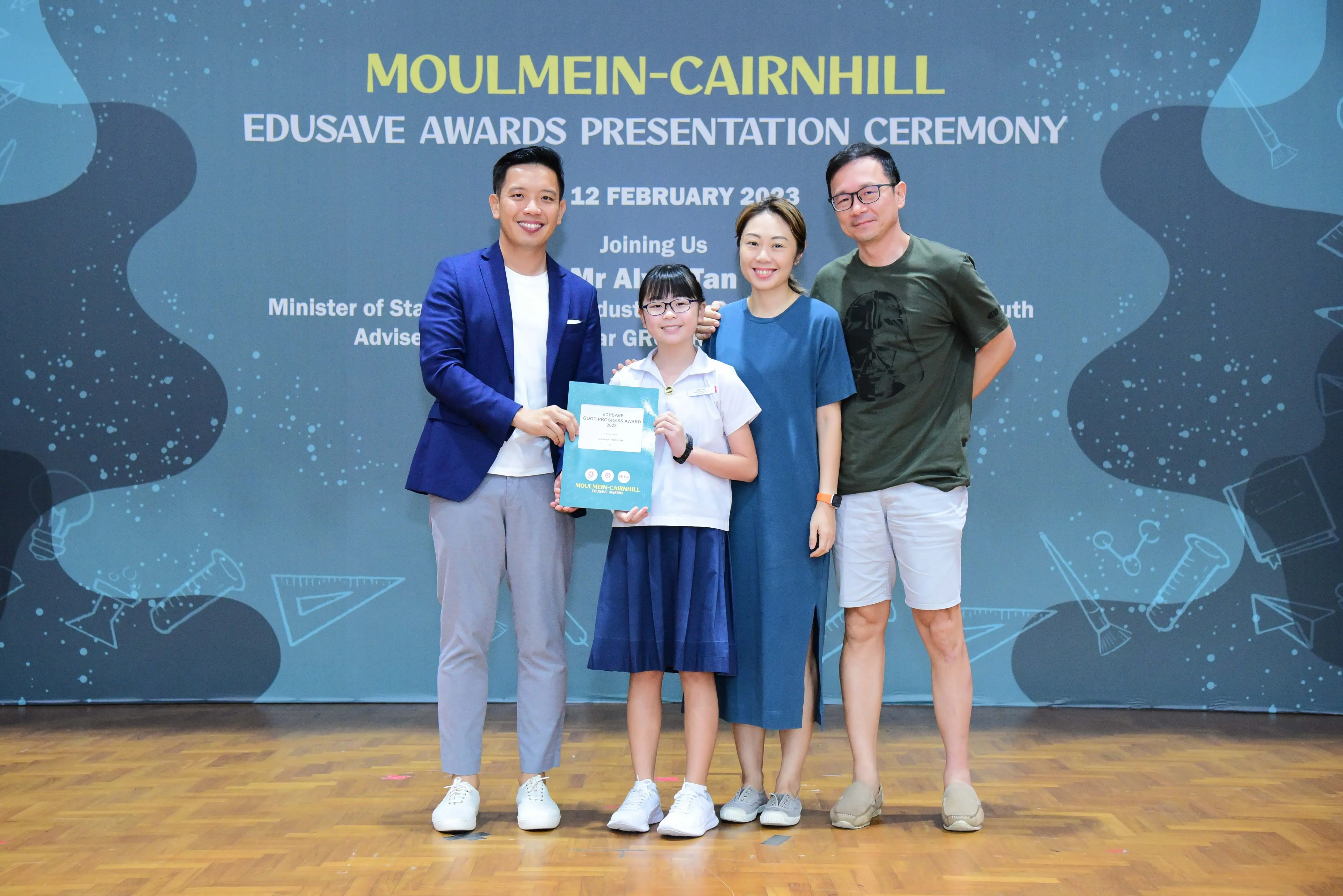 Four people pose at the Moulmein-Cairnhill Edusave Awards Presentation Ceremony, with a girl holding a certificate.