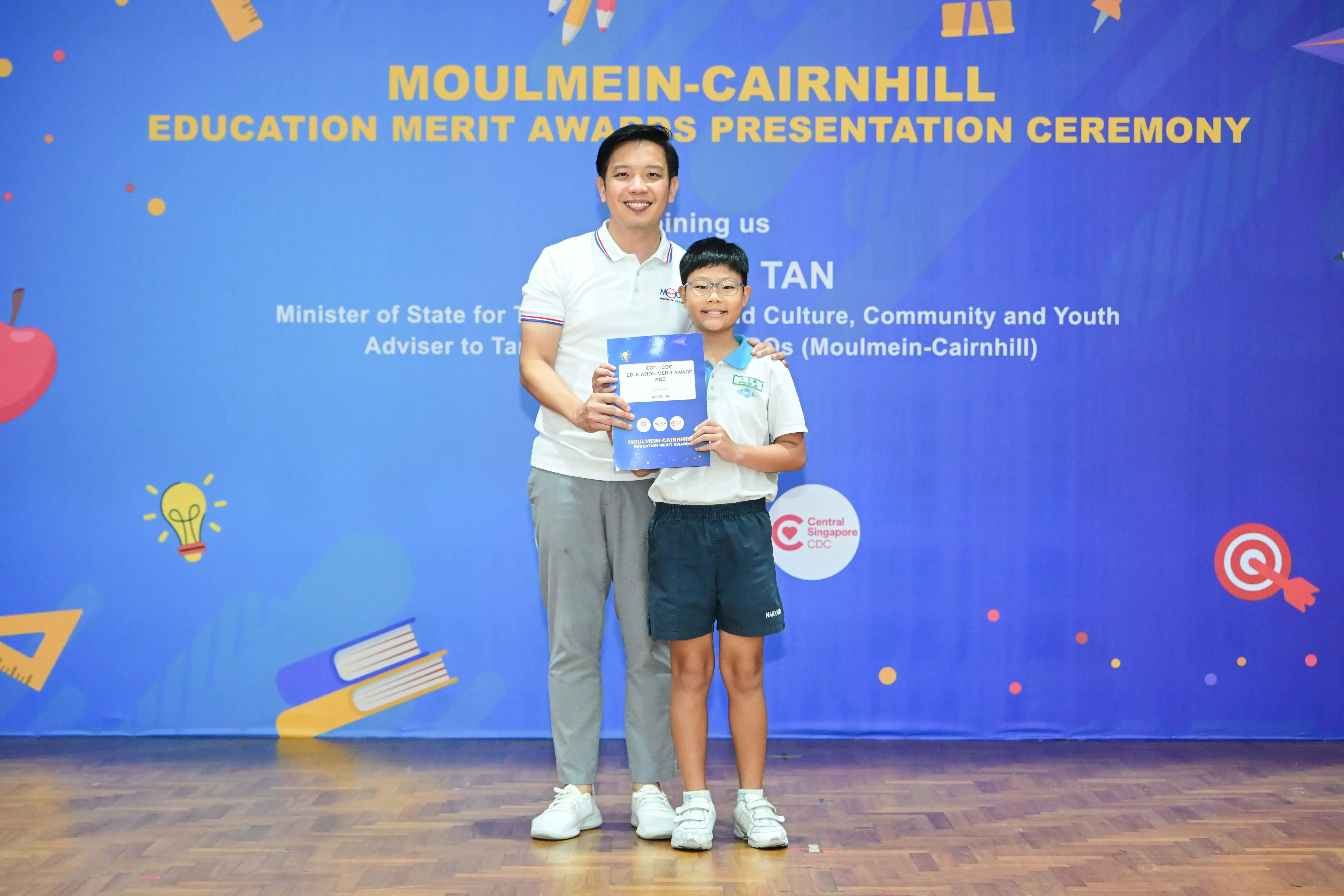 A man and a young boy holding an award certificate at the Moulmein-Cairnhill Education Merit Awards Ceremony.