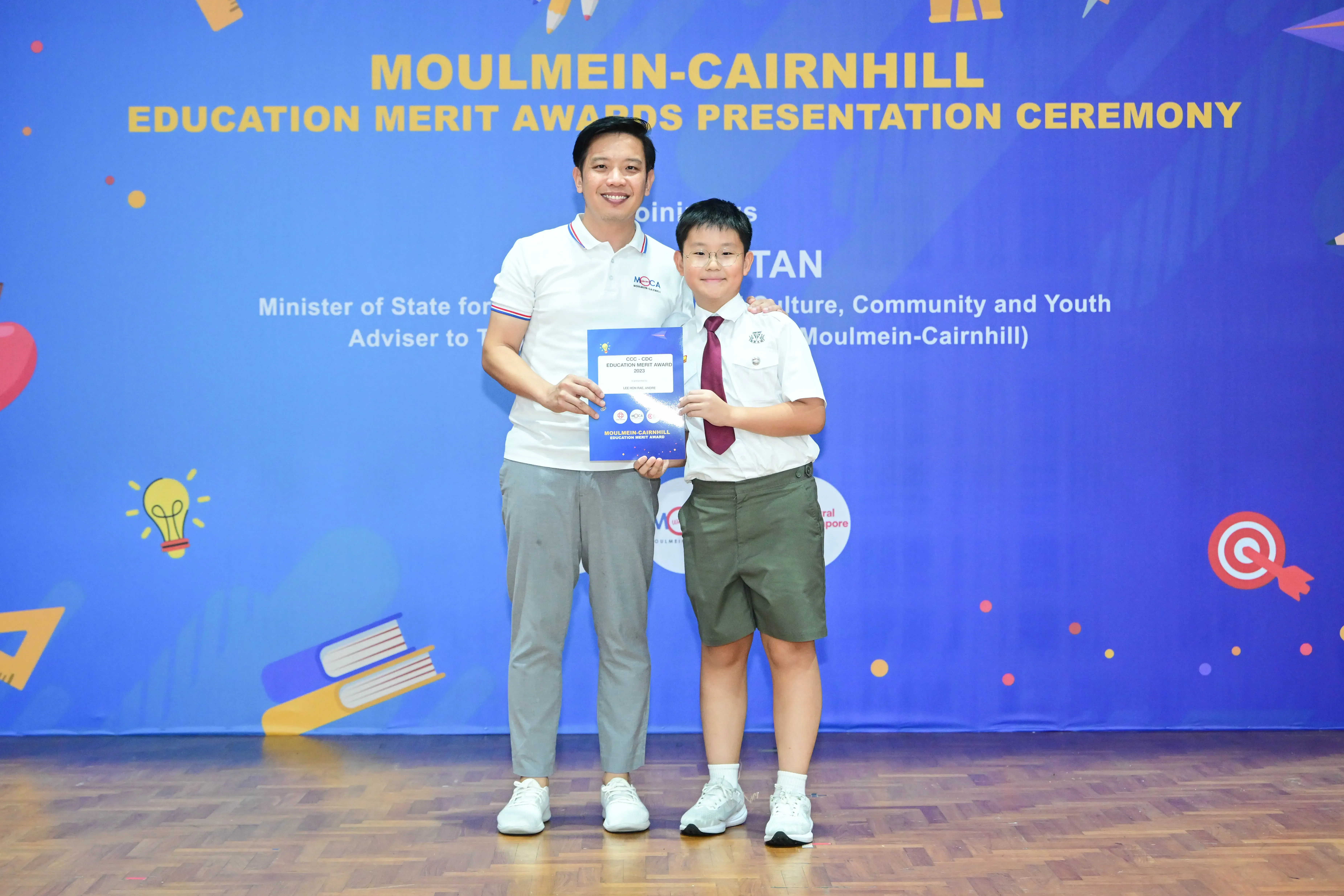 An adult and a young boy hold a certificate together at the Moulmein-Cairnhill Education Merit Awards ceremony.