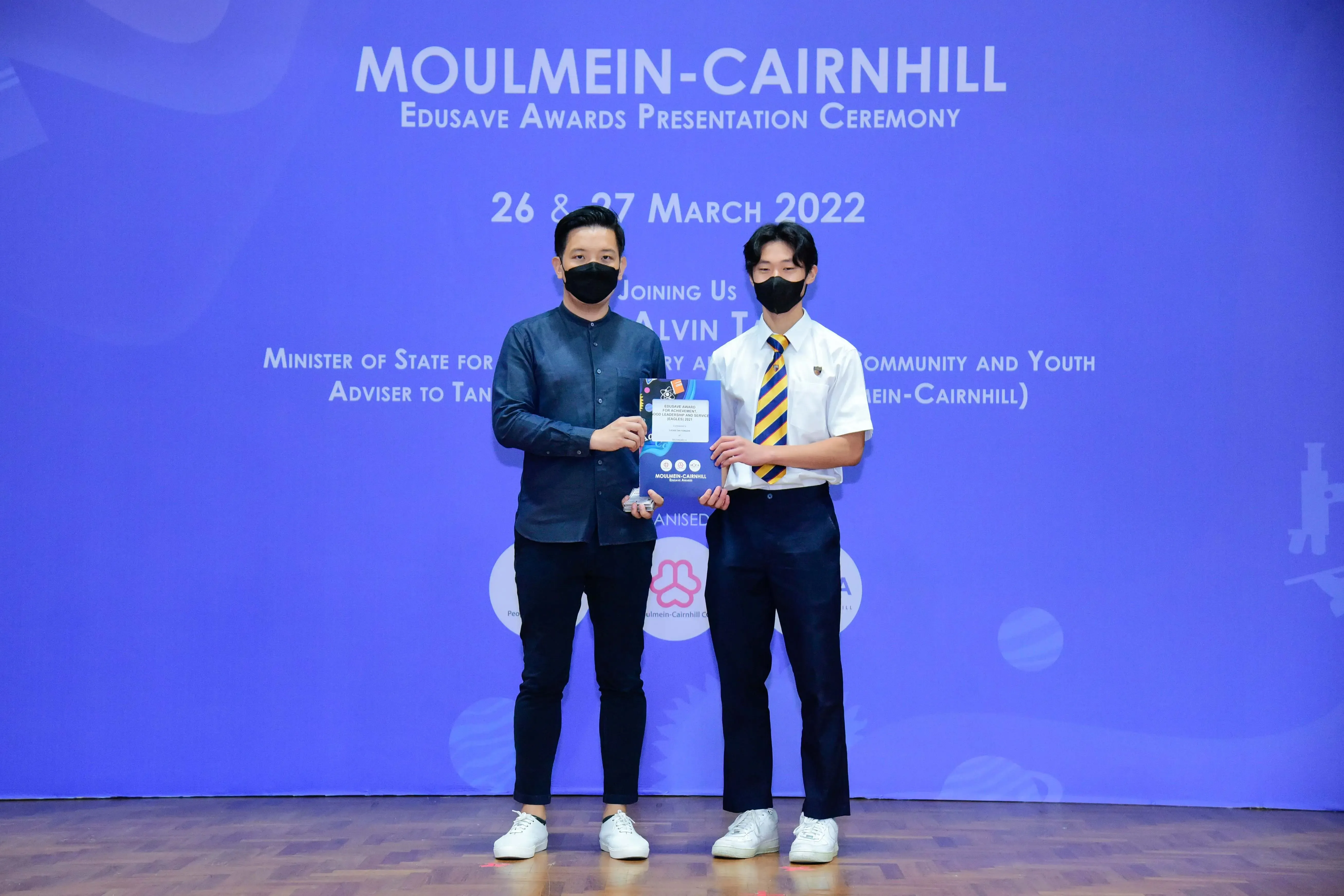 Two masked men hold an award in front of a blue Moulmein-Cairnhill Edusave Awards Ceremony backdrop dated March 2022.
