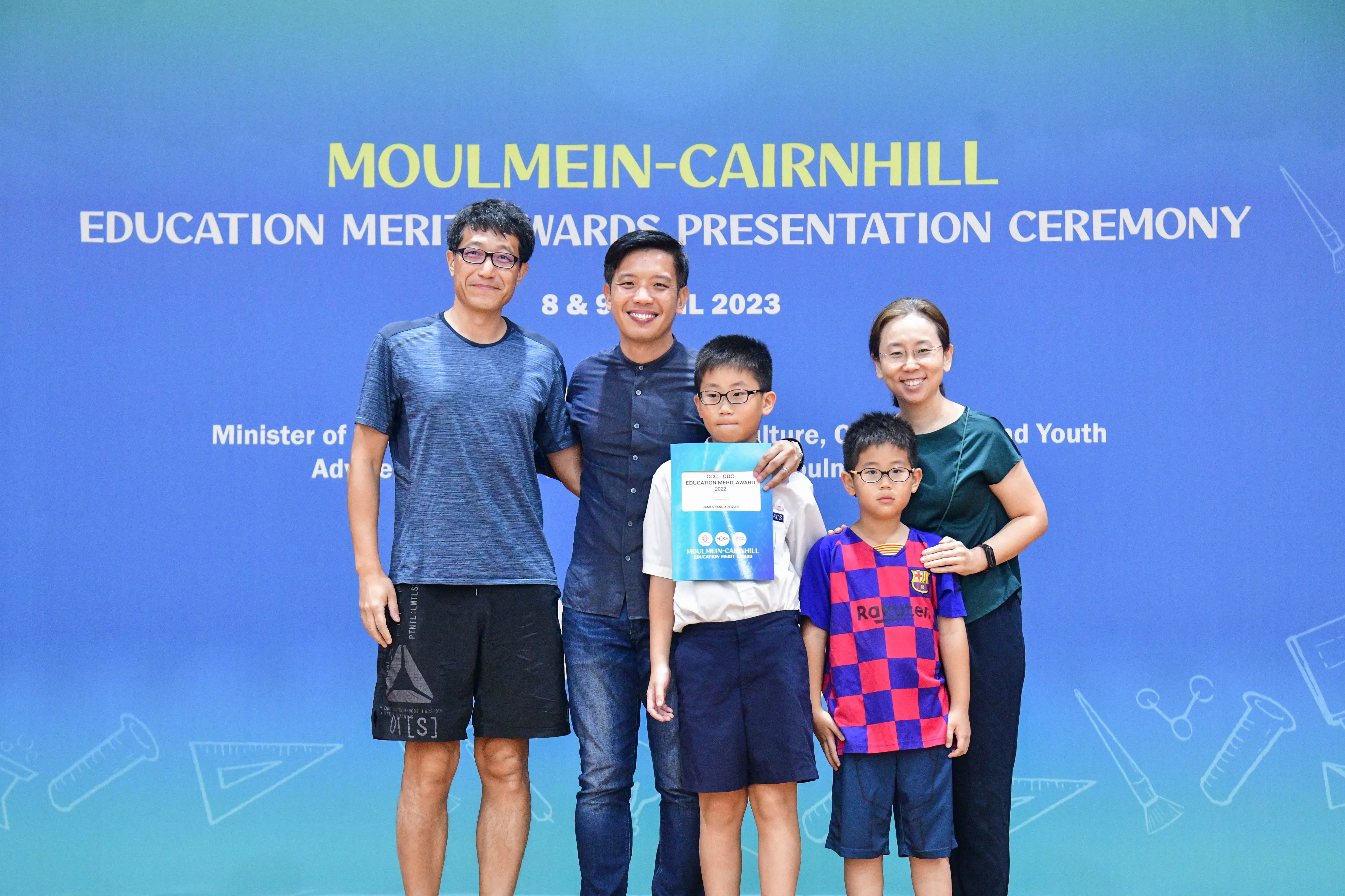 Five people pose before a Moulmein-Cairnhill Education Merit Awards Presentation Ceremony banner in 2023.