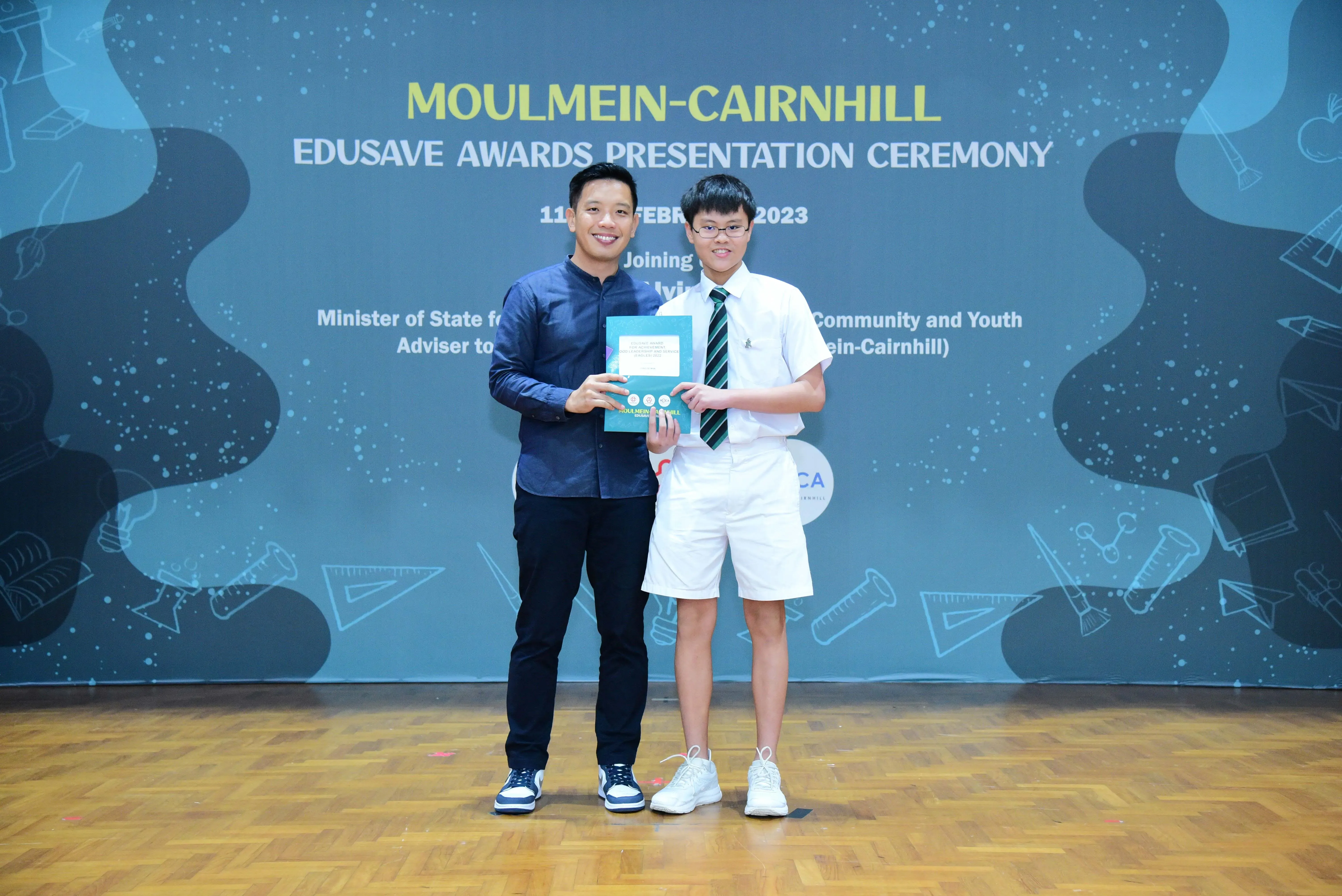 An adult and a student in school uniform hold an award certificate at the Moulmein-Cairnhill Edusave Awards Ceremony 2023.