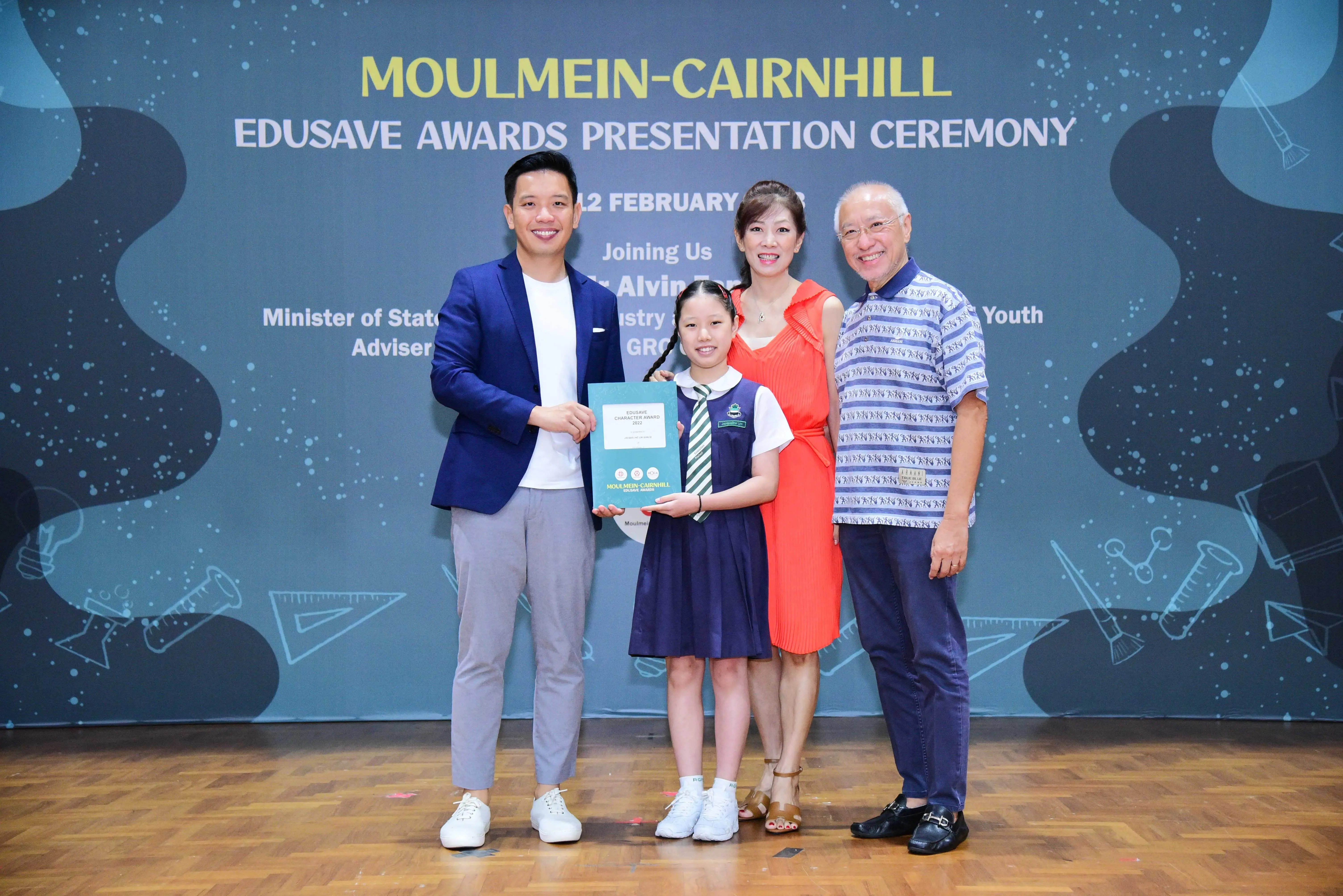 Four people pose at the Moulmein-Cairnhill Edusave Awards Presentation Ceremony, with a girl holding an award certificate.