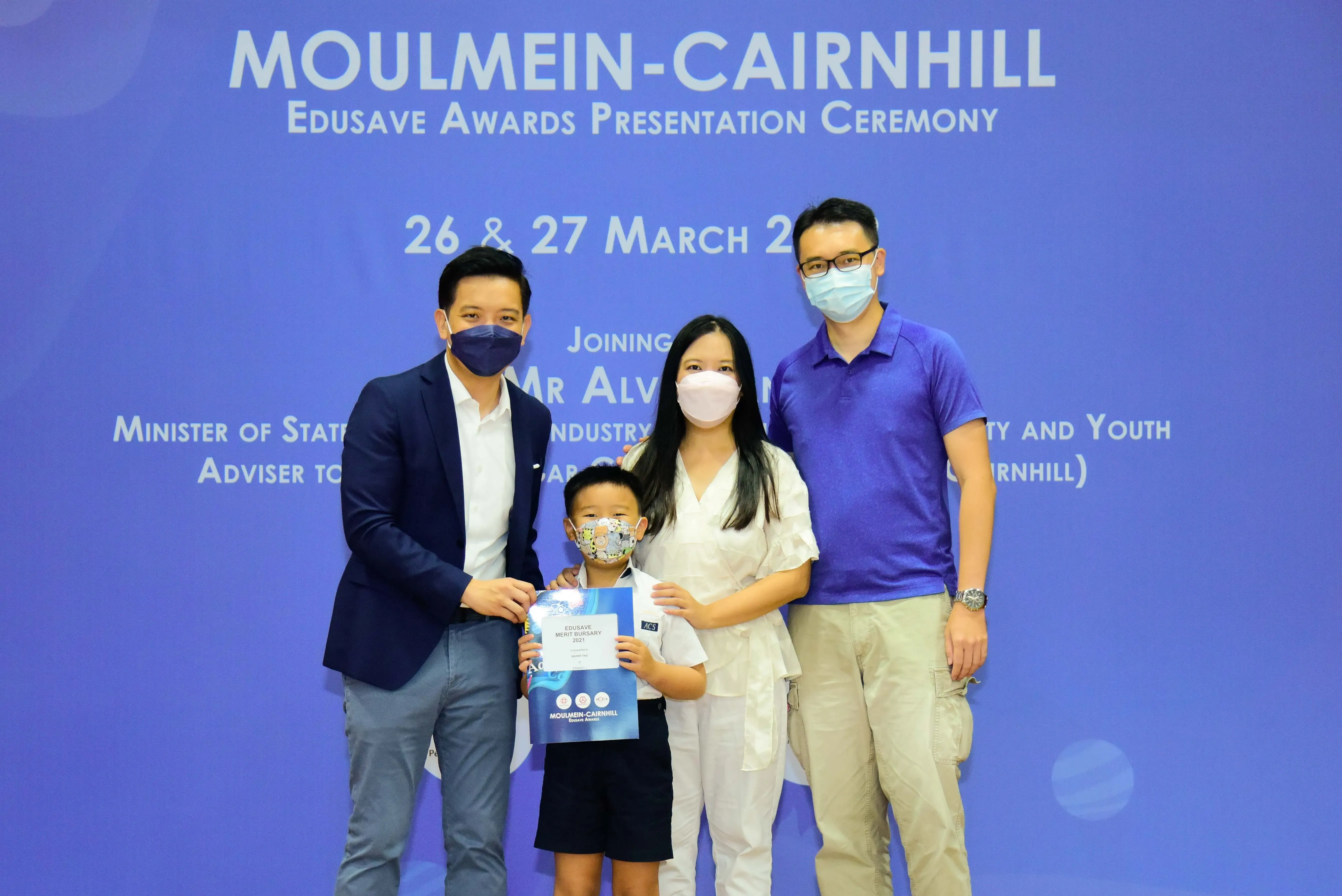 Four masked people pose at the Moulmein-Cairnhill Edusave Awards Presentation Ceremony, with a child holding a certificate.