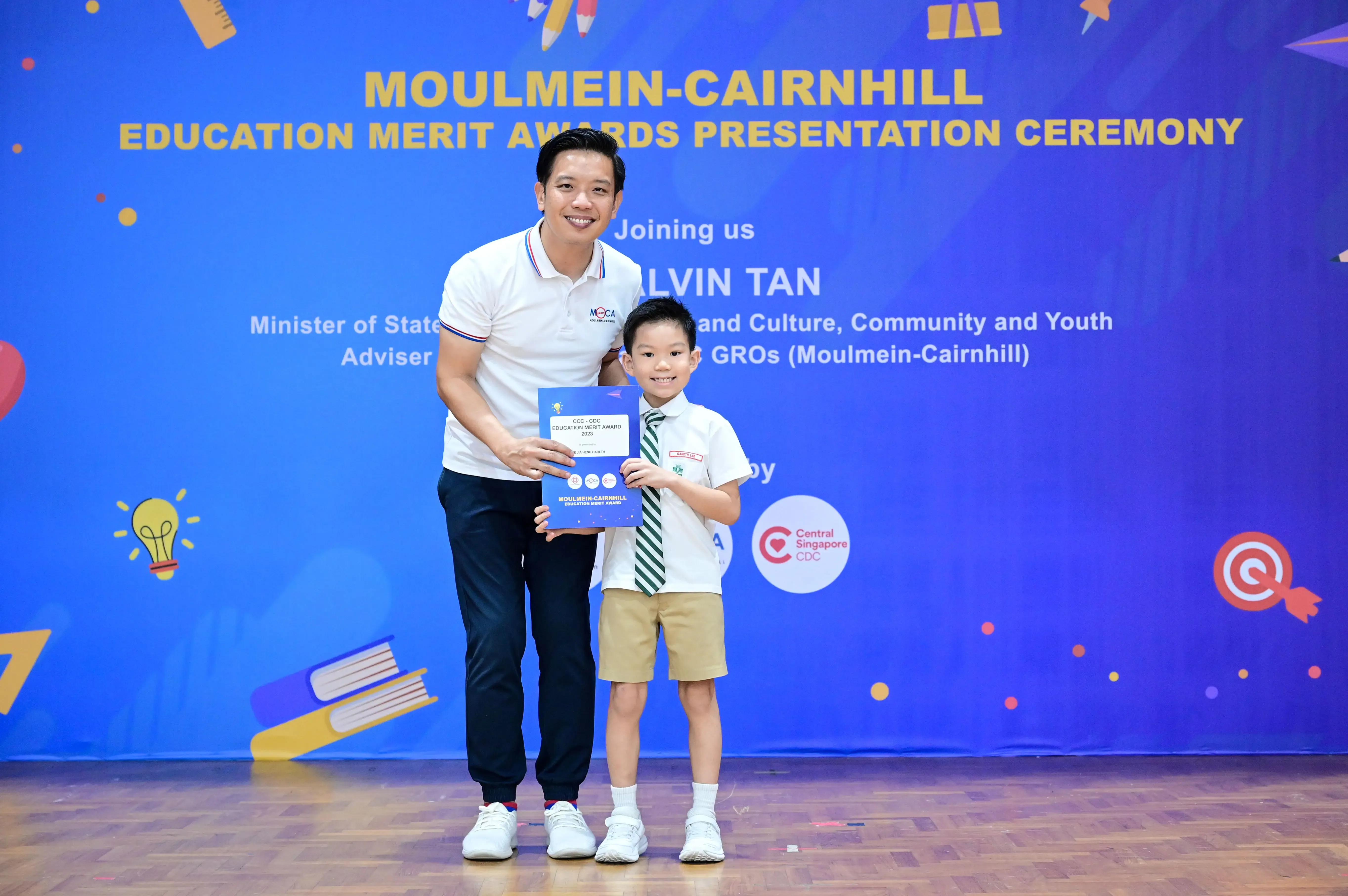 Man and young boy in school uniform hold an award certificate at Moulmein-Cairnhill Education Merit Awards ceremony.