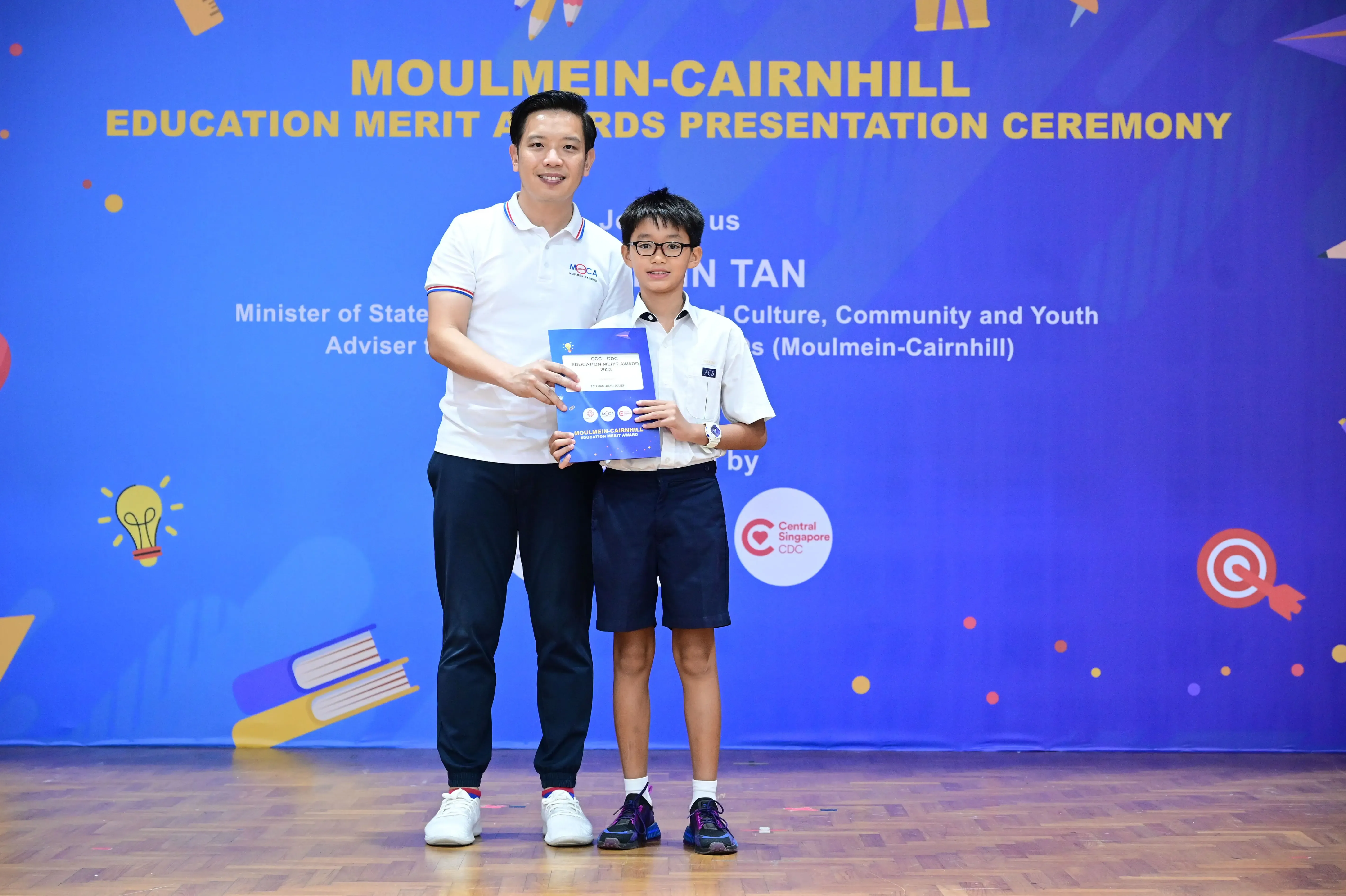 An adult and a young student hold an award certificate at the Moulmein-Cairnhill Education Merit Awards Presentation Ceremony.