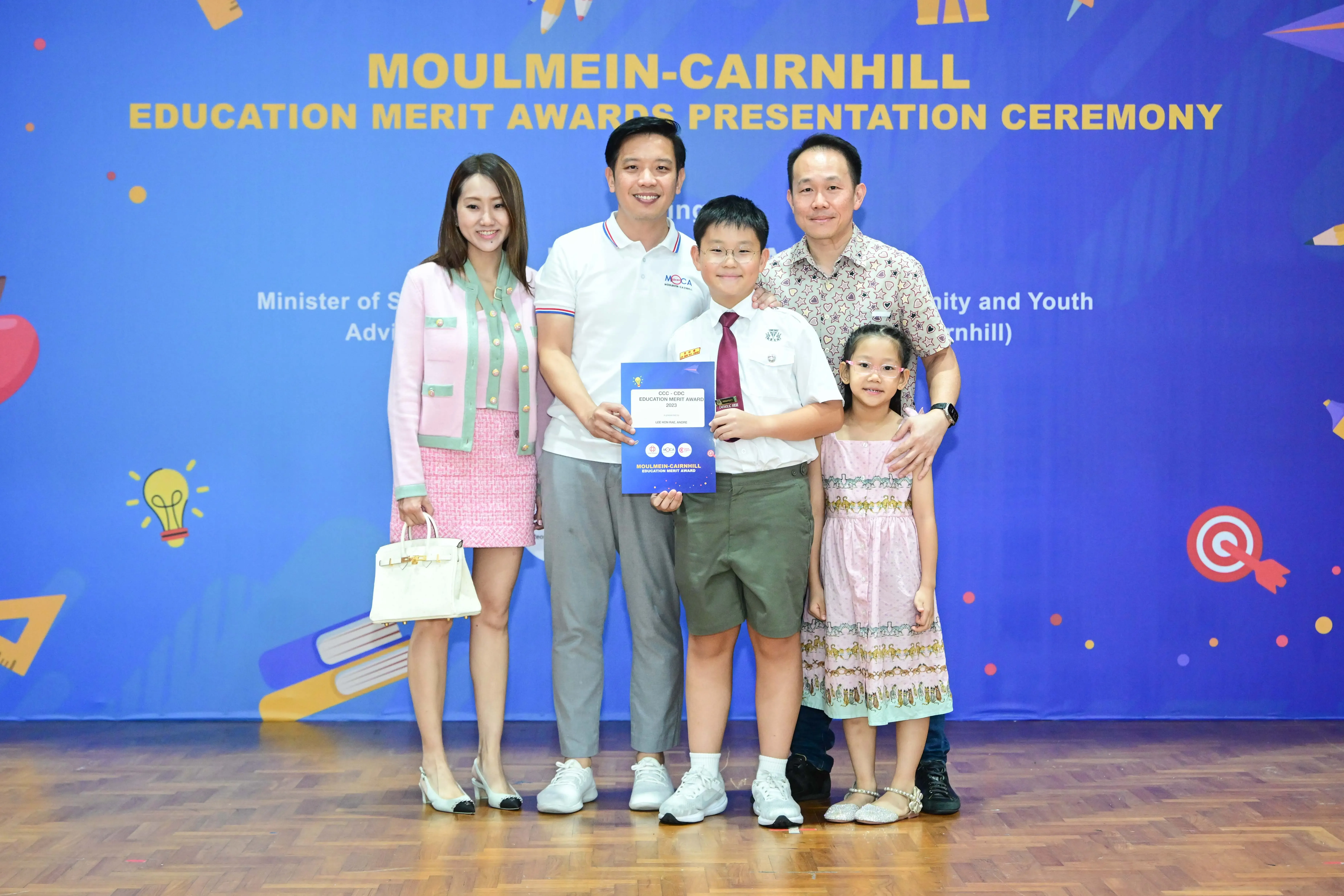 Five people pose before a Moulmein-Cairnhill Education Merit Awards banner, with a boy holding an award certificate.
