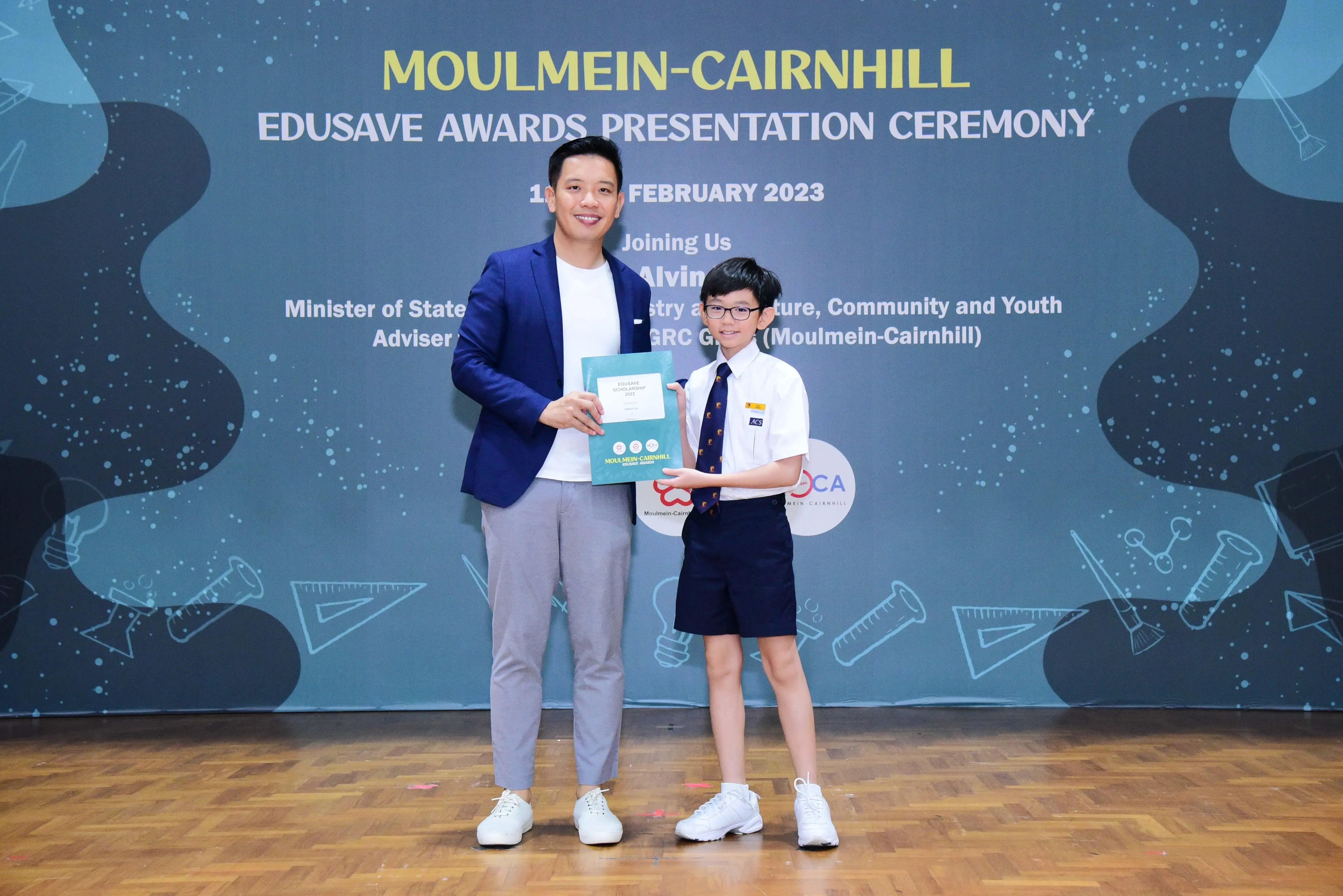 An adult in a blue blazer and a young student in school uniform hold a certificate at the Moulmein-Cairnhill Edusave Awards 2023.