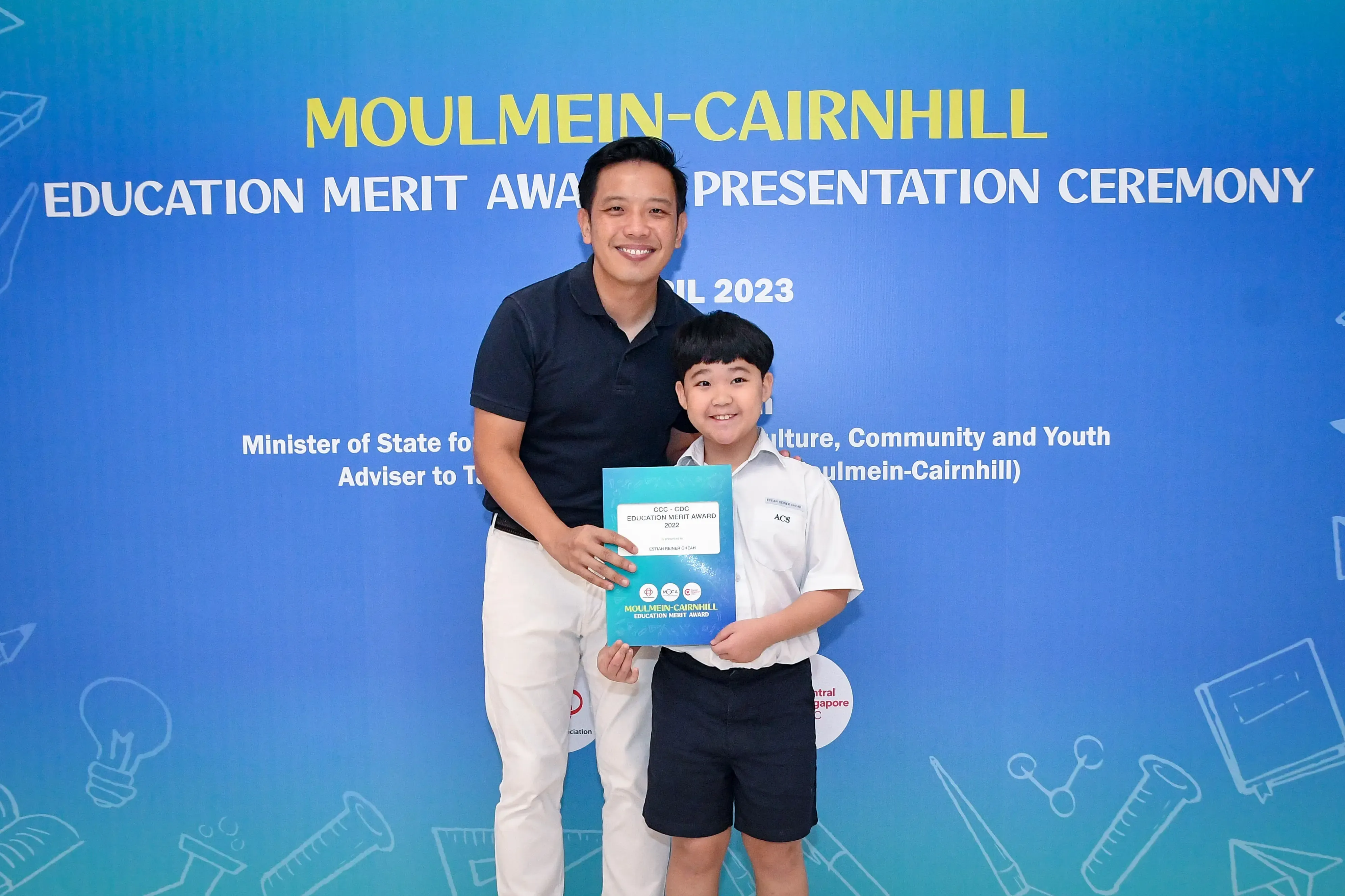 A man and a young boy smile holding an award certificate at the 2023 Moulmein-Cairnhill Education Merit Award ceremony.