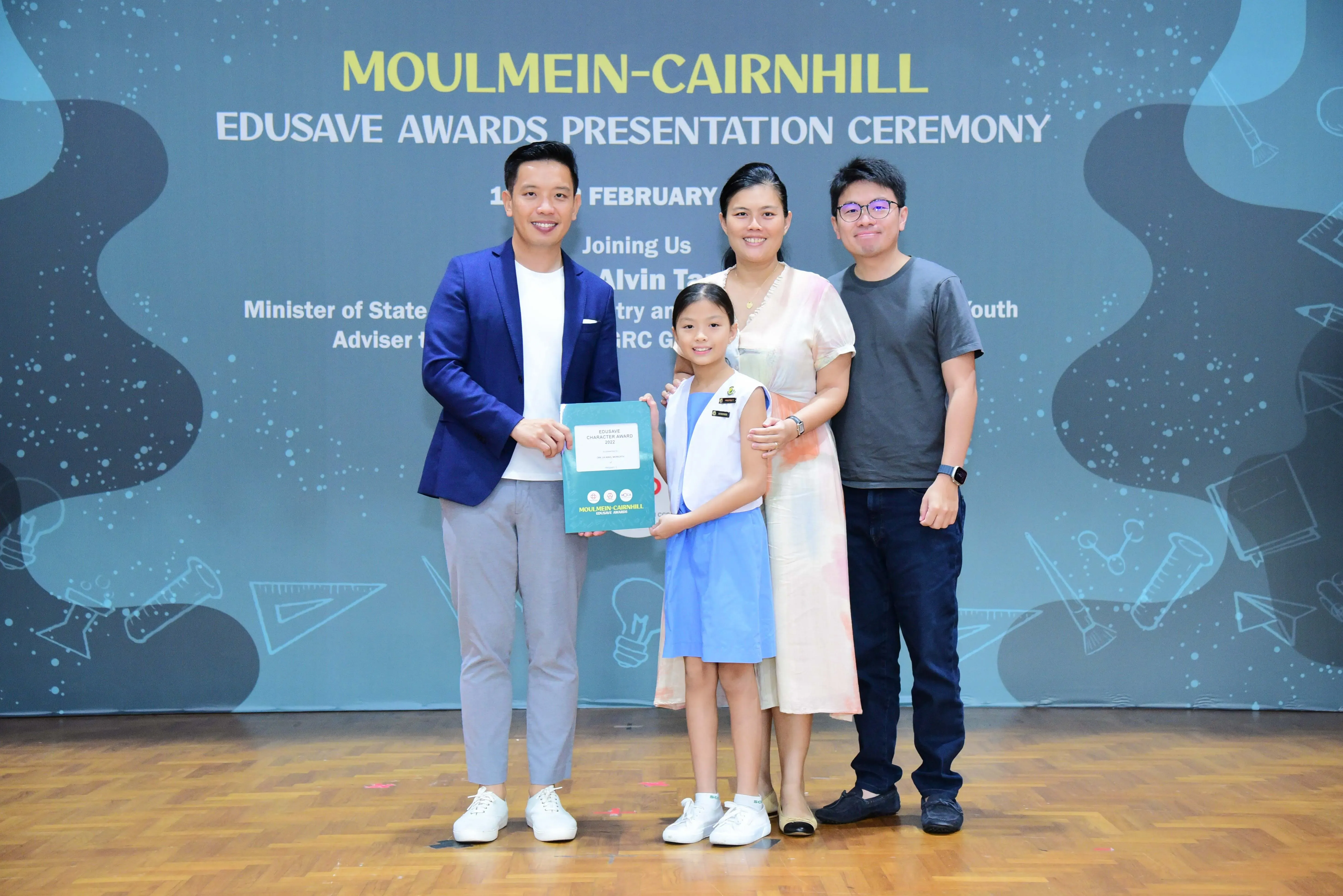 Four people pose with an award certificate at the Moulmein-Cairnhill Edusave Awards Presentation Ceremony backdrop.
