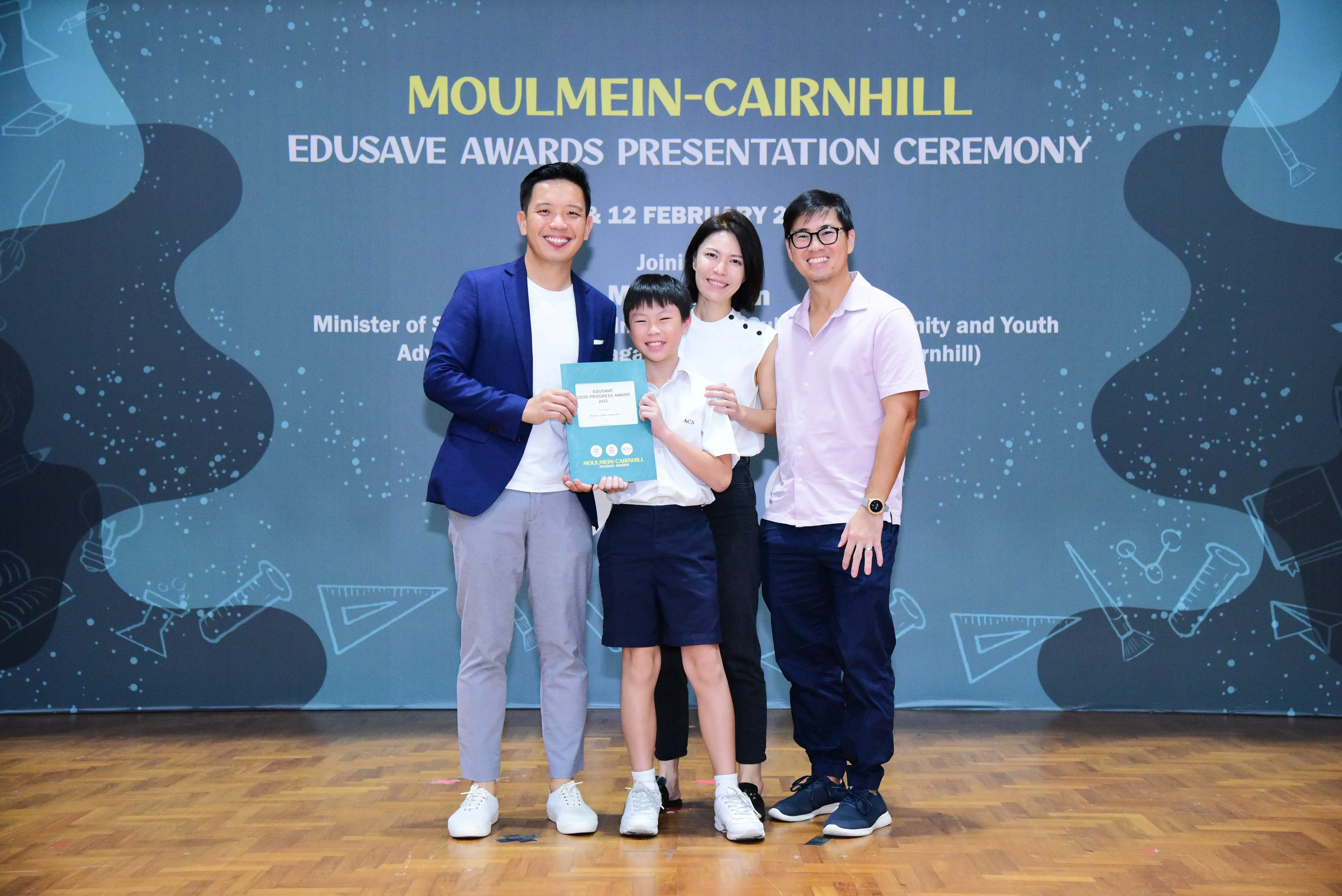 Four people pose smiling at the Moulmein-Cairnhill Edusave Awards Presentation Ceremony, with a child holding a certificate.