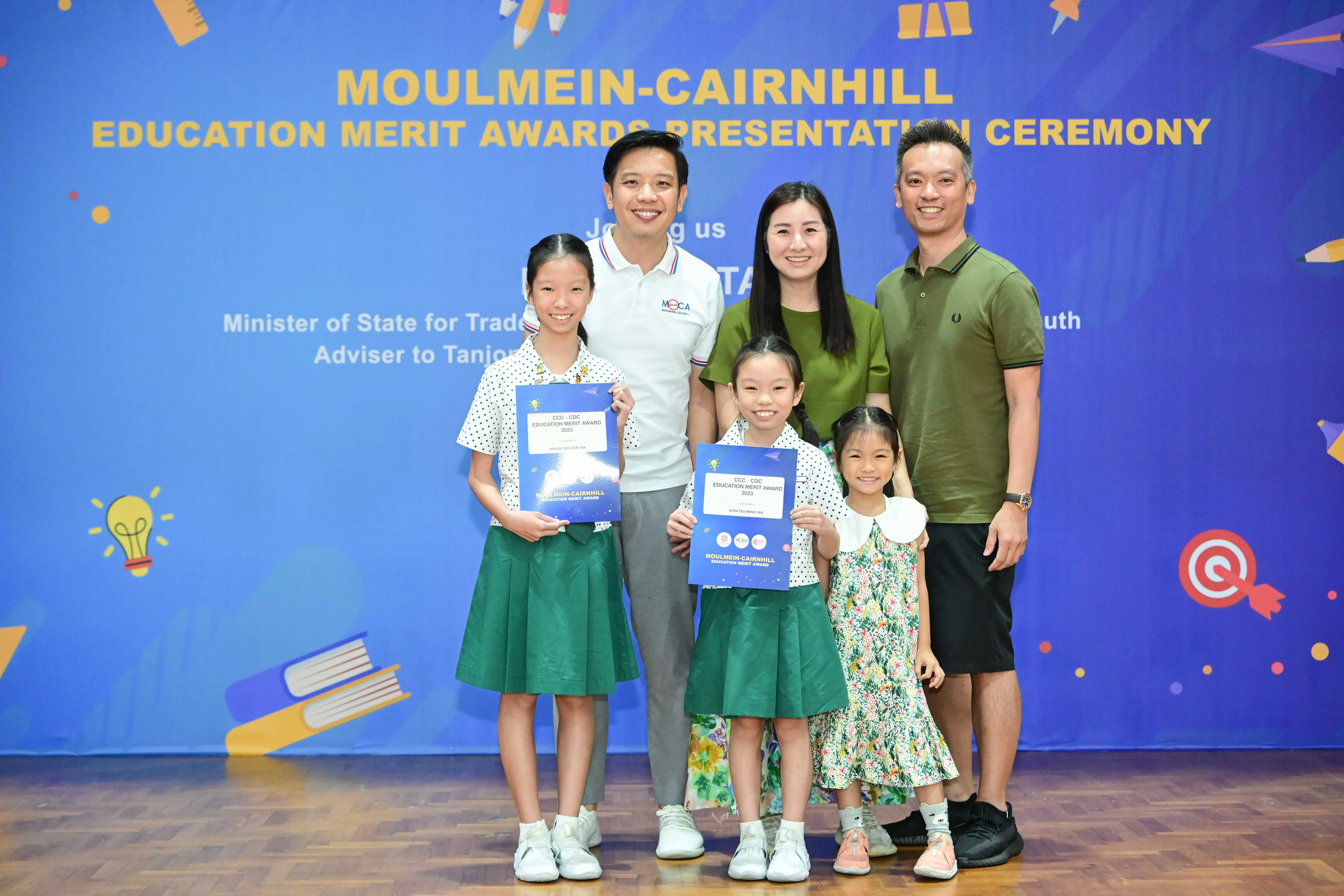 Three adults and three young girls pose at the Moulmein-Cairnhill Education Merit Awards, two girls holding award certificates.