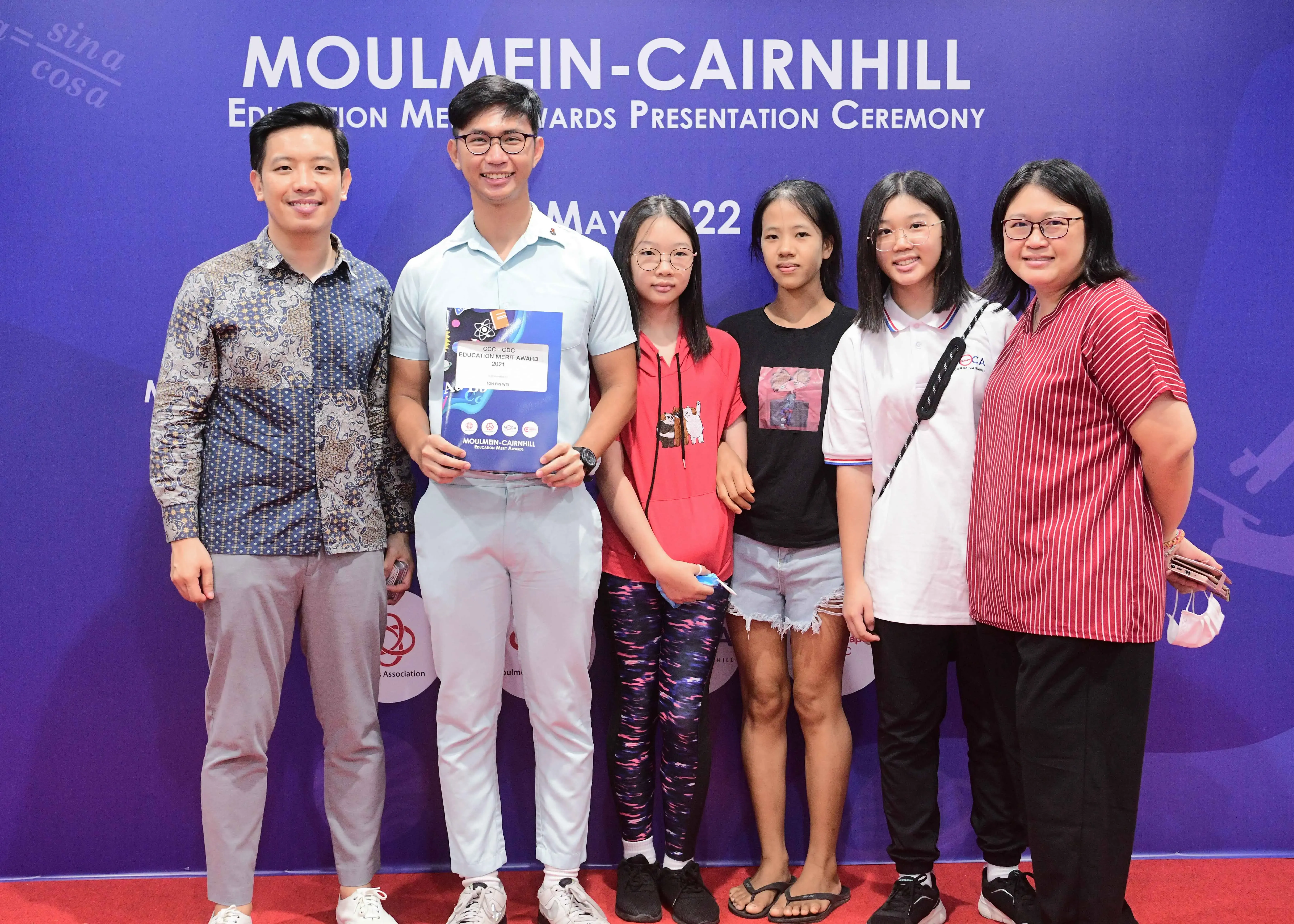 Six people pose smiling before a blue Moulmein-Cairnhill Education Merit Awards Presentation Ceremony banner dated May 2022.