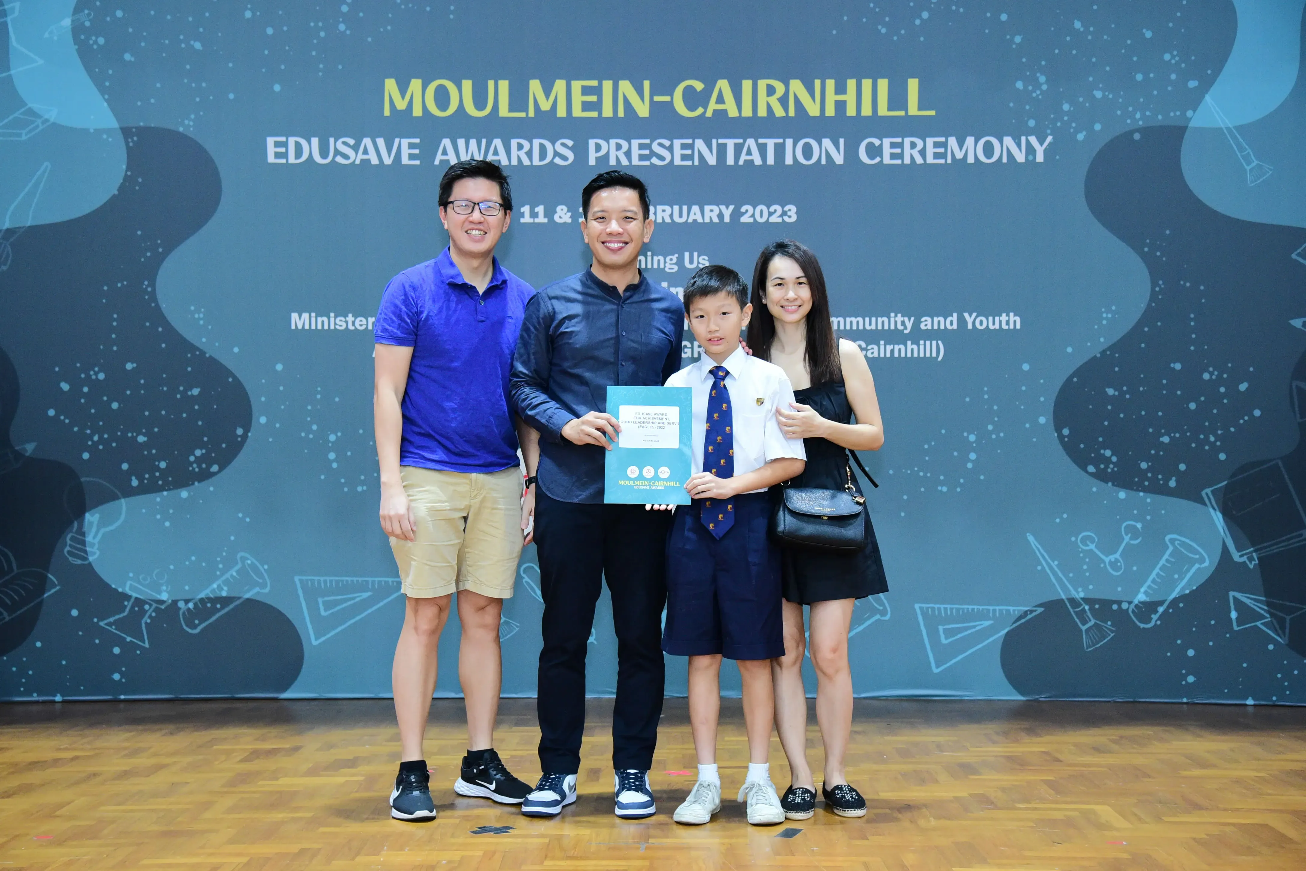 Three adults and a boy in school uniform hold a certificate at the Moulmein-Cairnhill Edusave Awards Ceremony 2023.