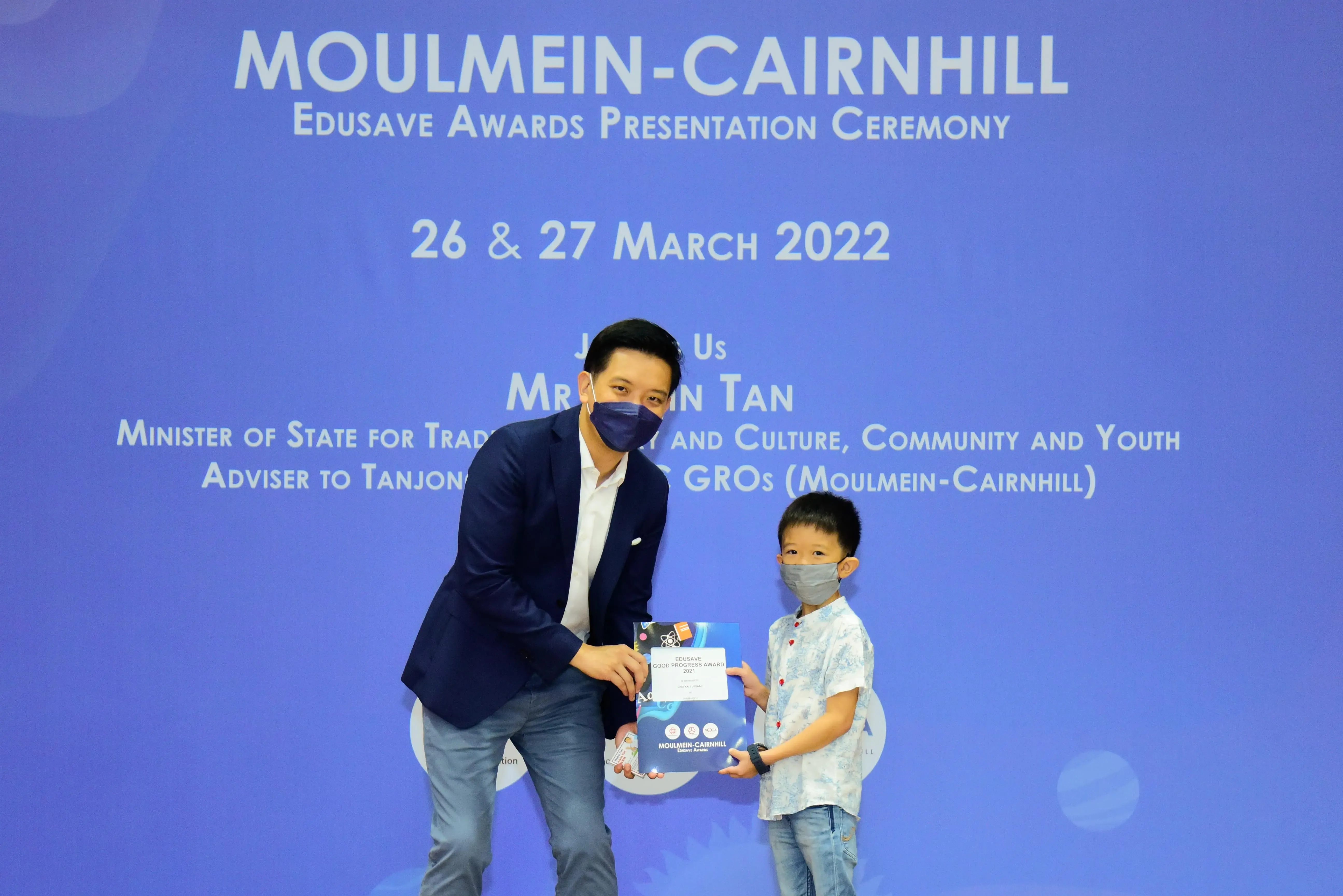 A masked man in a blazer presents an award to a masked young boy at the Moulmein-Cairnhill Edusave Awards Ceremony 2022.