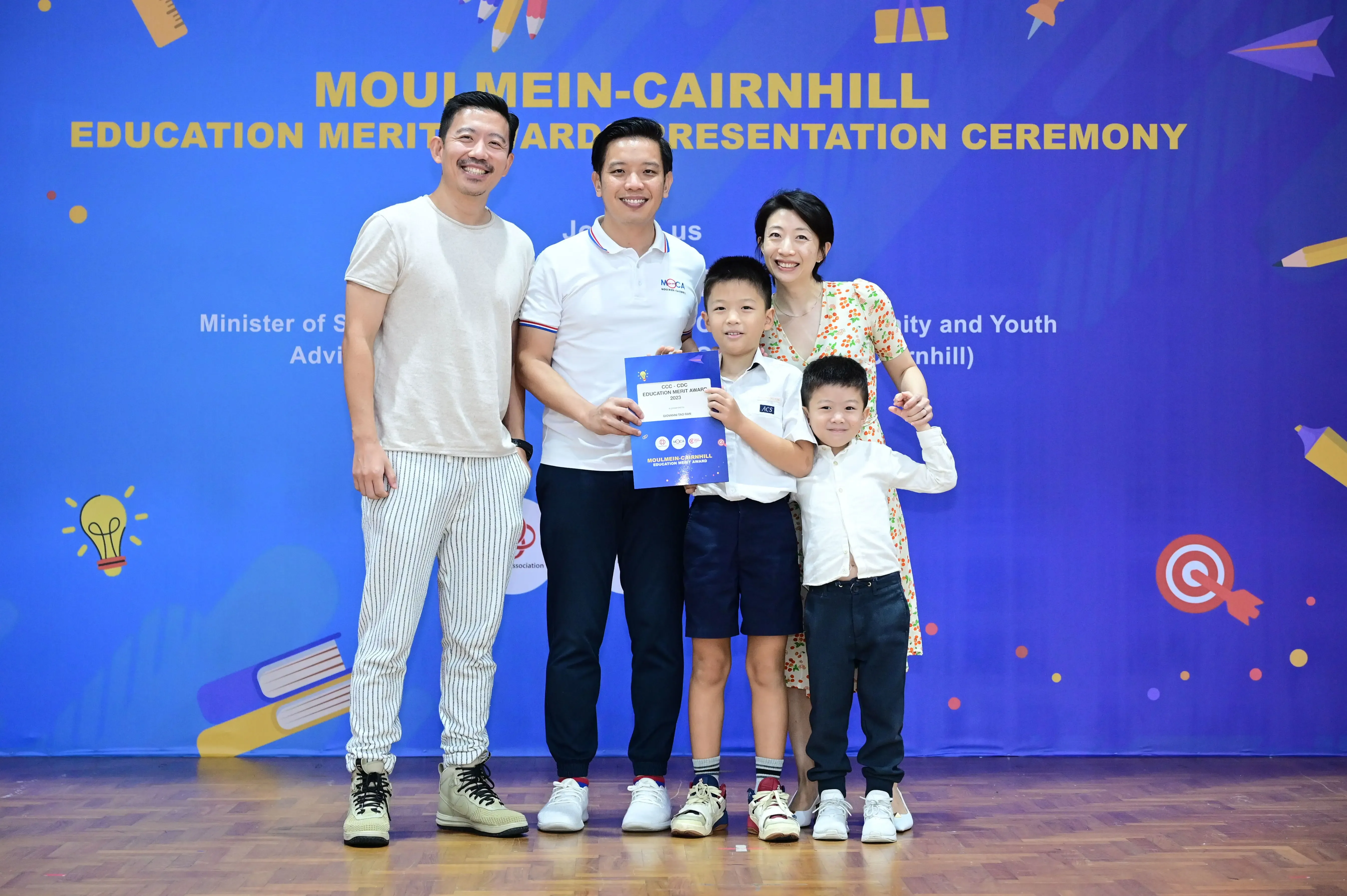 Five people pose smiling at the Moulmein-Cairnhill Education Merit Award Presentation Ceremony, a boy holding a certificate.