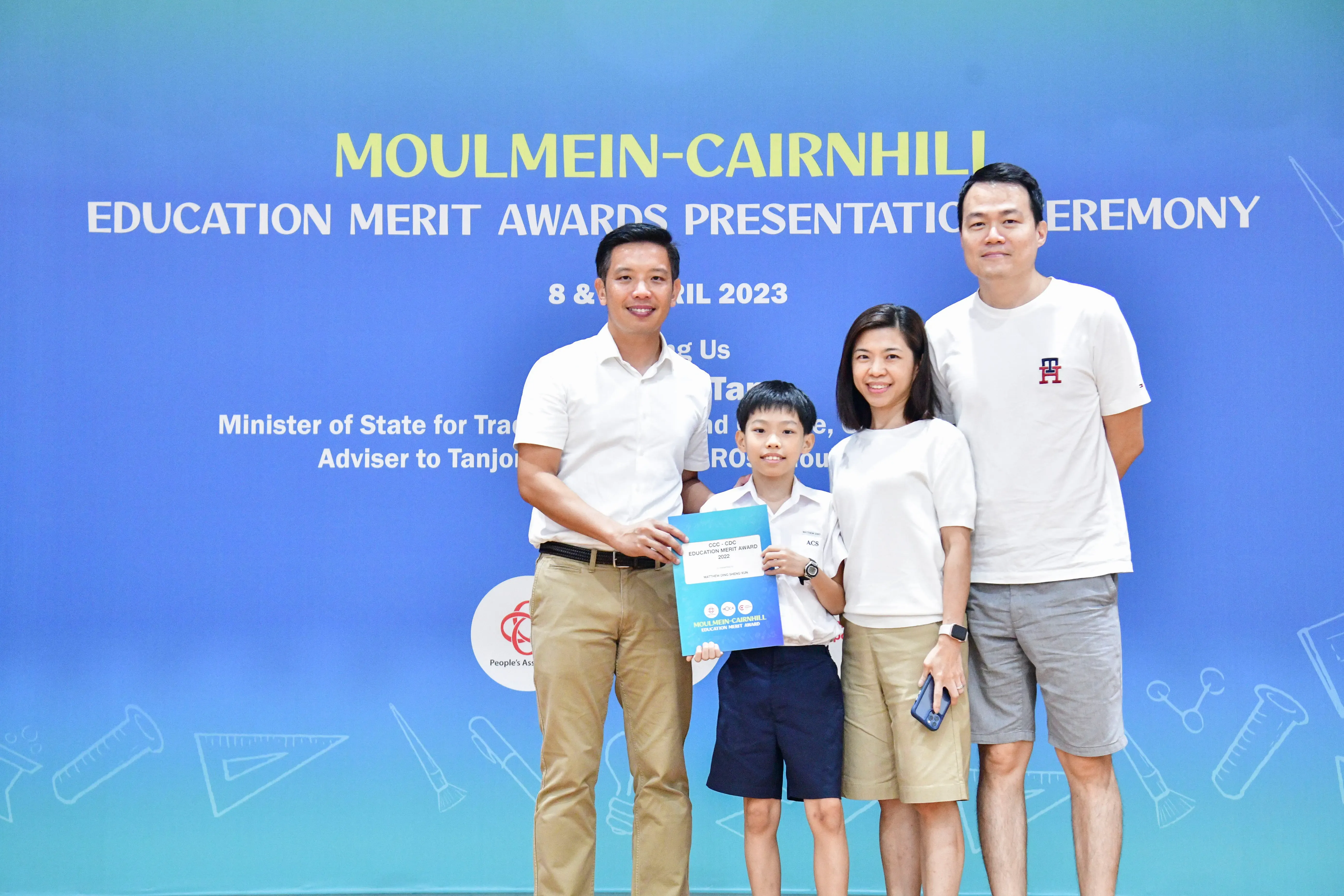 Four people pose before a Moulmein-Cairnhill Education Merit Awards 2023 banner, with a boy holding a certificate.