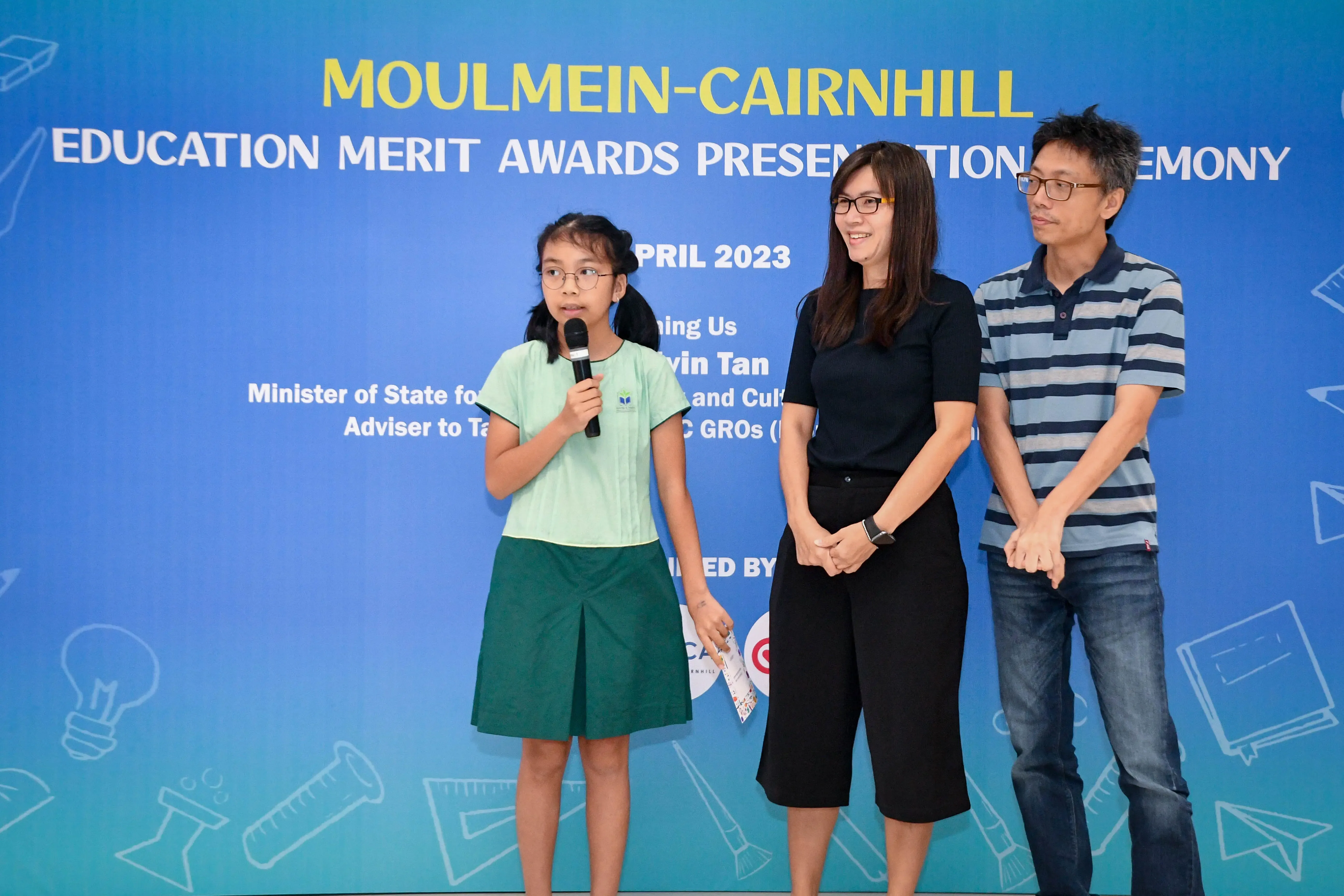 A girl in school uniform speaks into a microphone beside two adults at Moulmein-Cairnhill Education Merit Awards 2023.