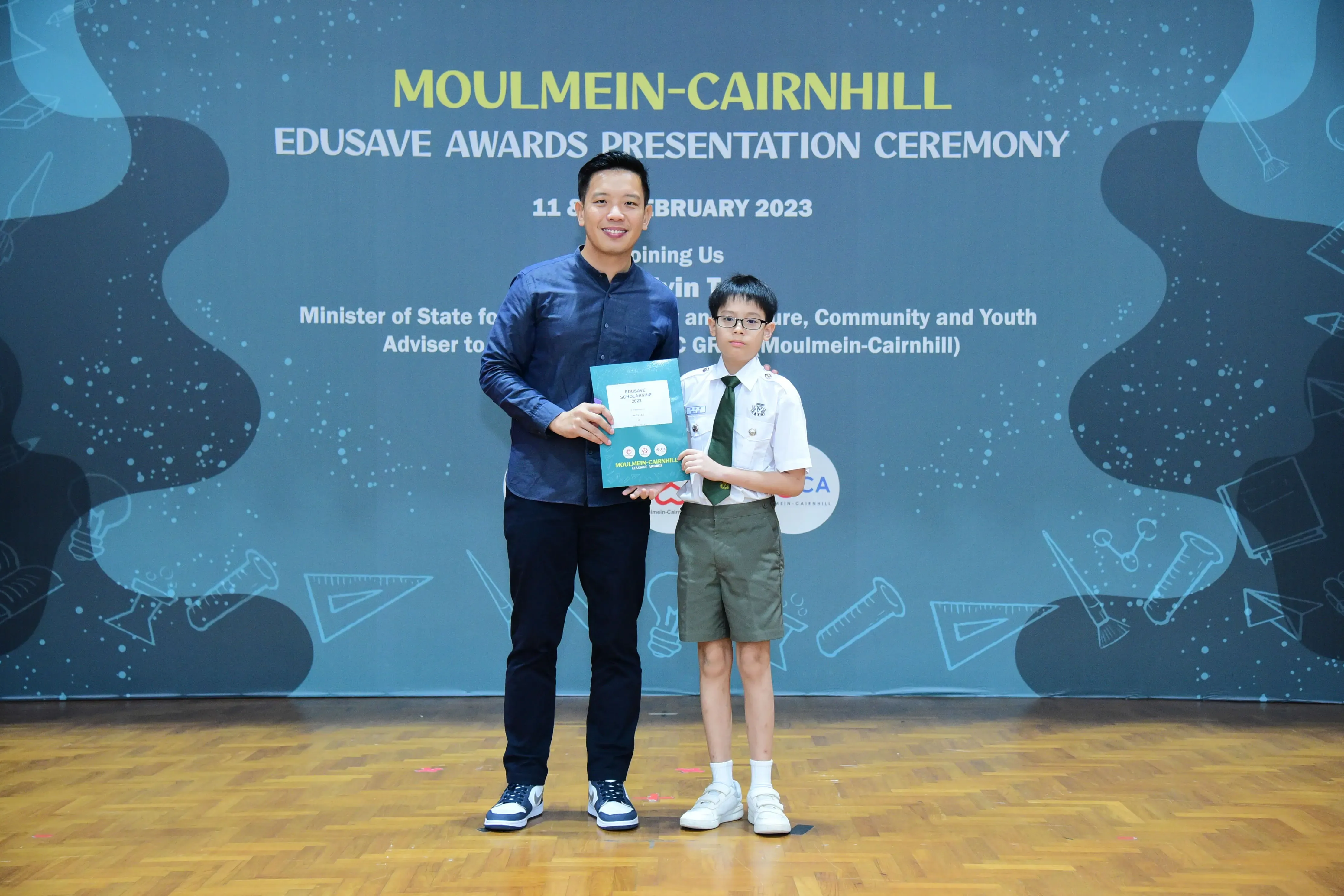 An adult and a uniformed boy hold a certificate at the Moulmein-Cairnhill Edusave Awards Presentation Ceremony 2023.