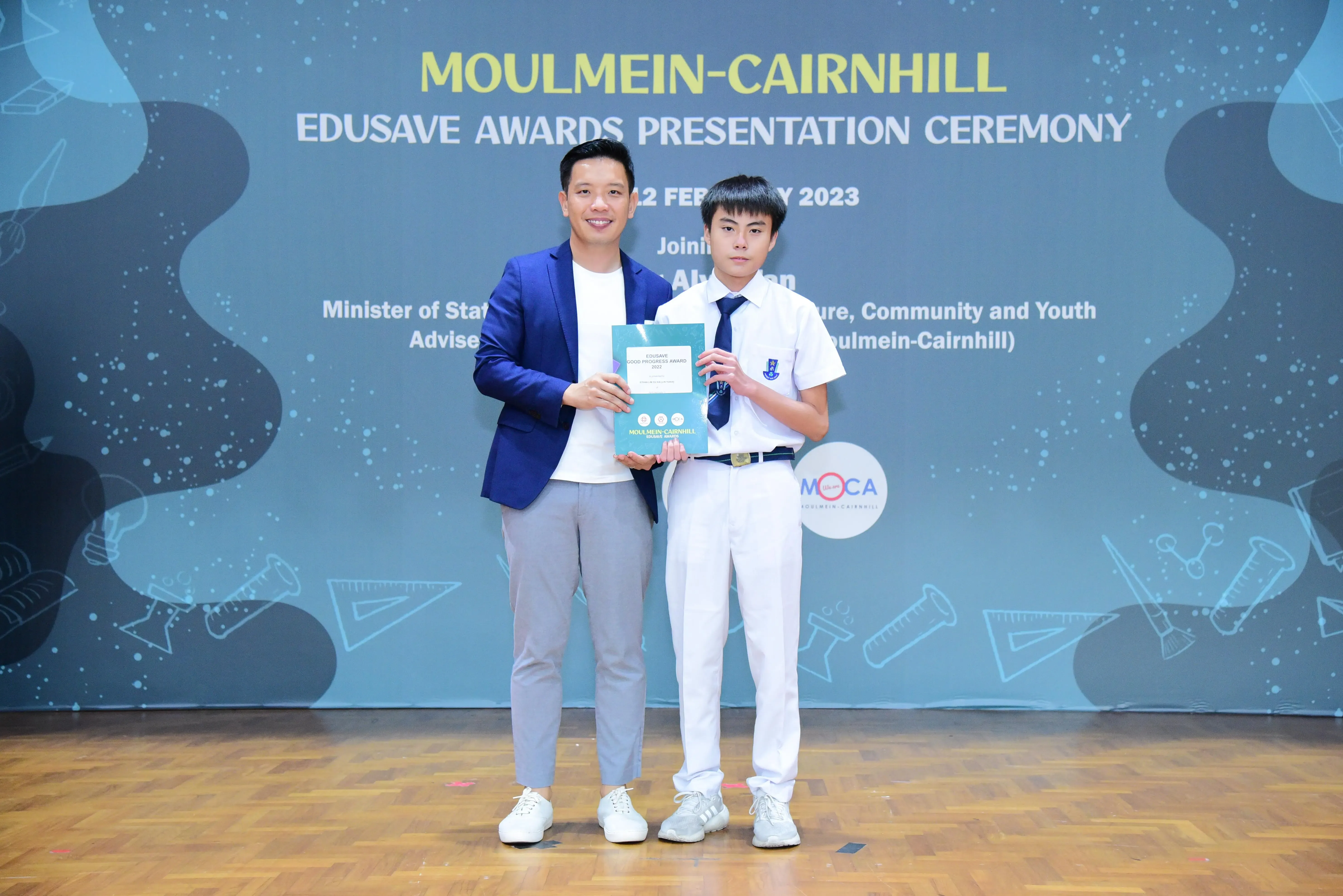 Two people hold an award certificate on stage at the Moulmein-Cairnhill Edusave Awards Presentation Ceremony 2023.
