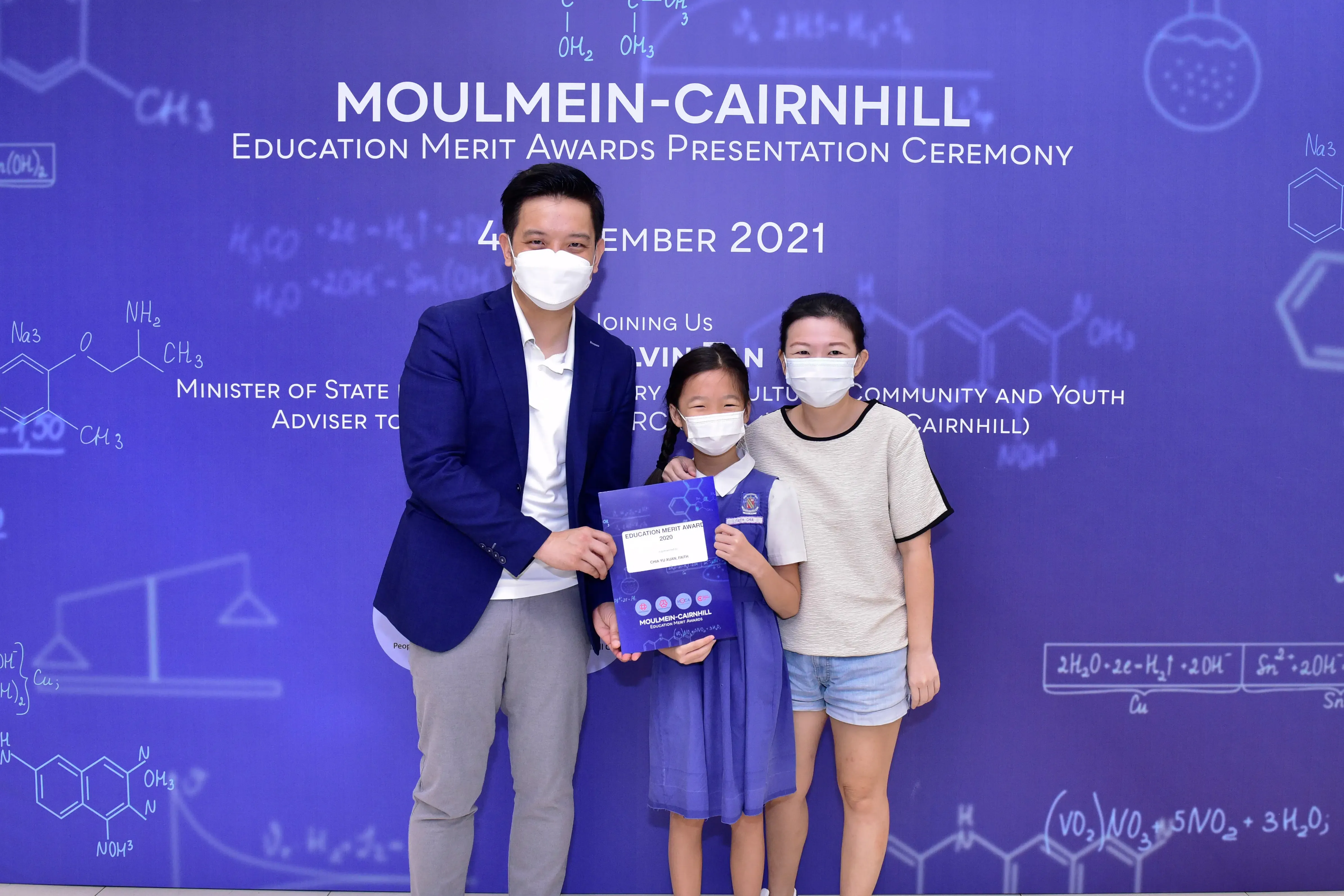 Three masked people pose at the Moulmein-Cairnhill Education Merit Awards Ceremony 2021, child holding a certificate.