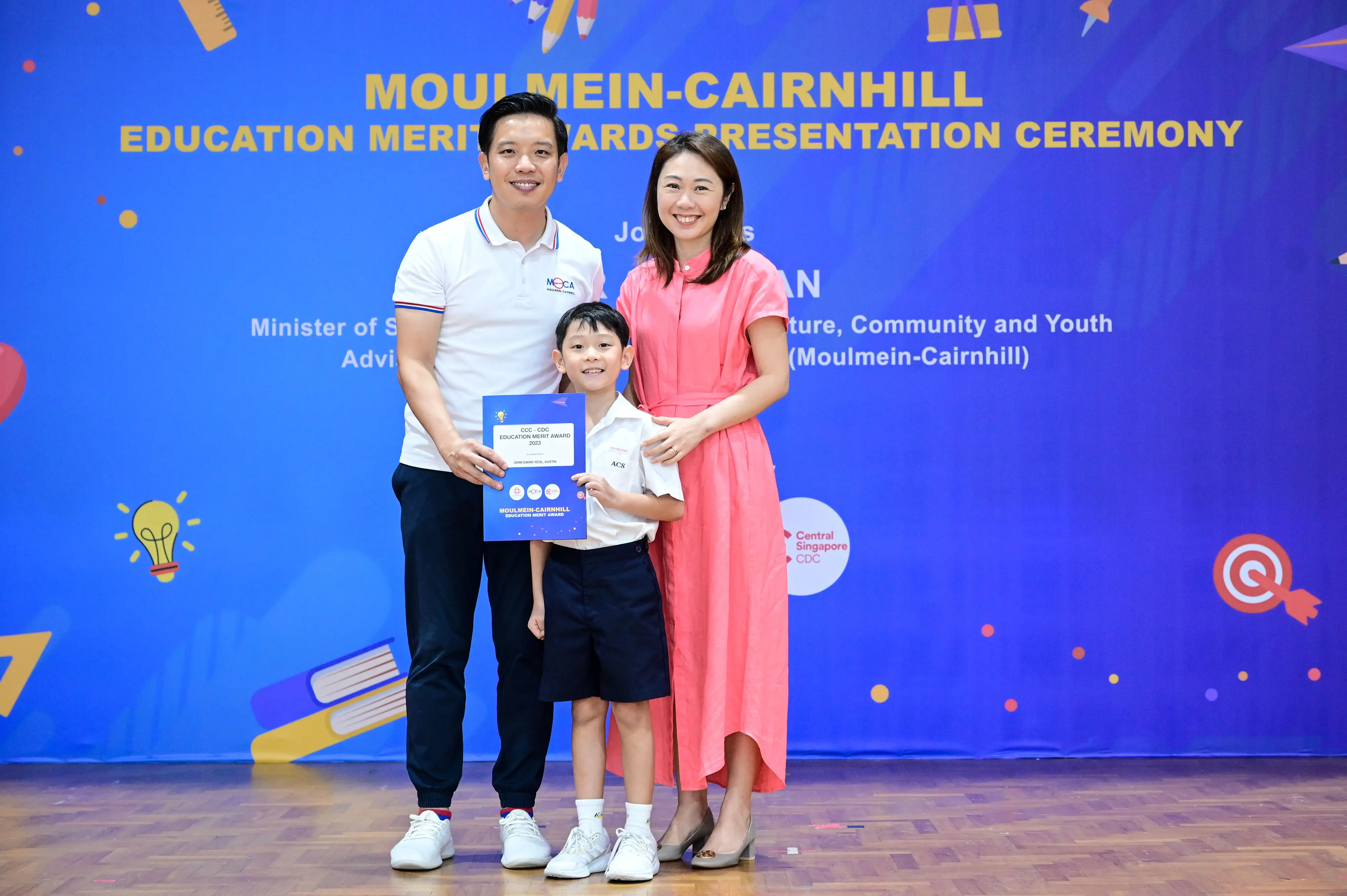 Two adults and a young boy hold a merit award certificate at the Moulmein-Cairnhill Education Merit Awards Ceremony.