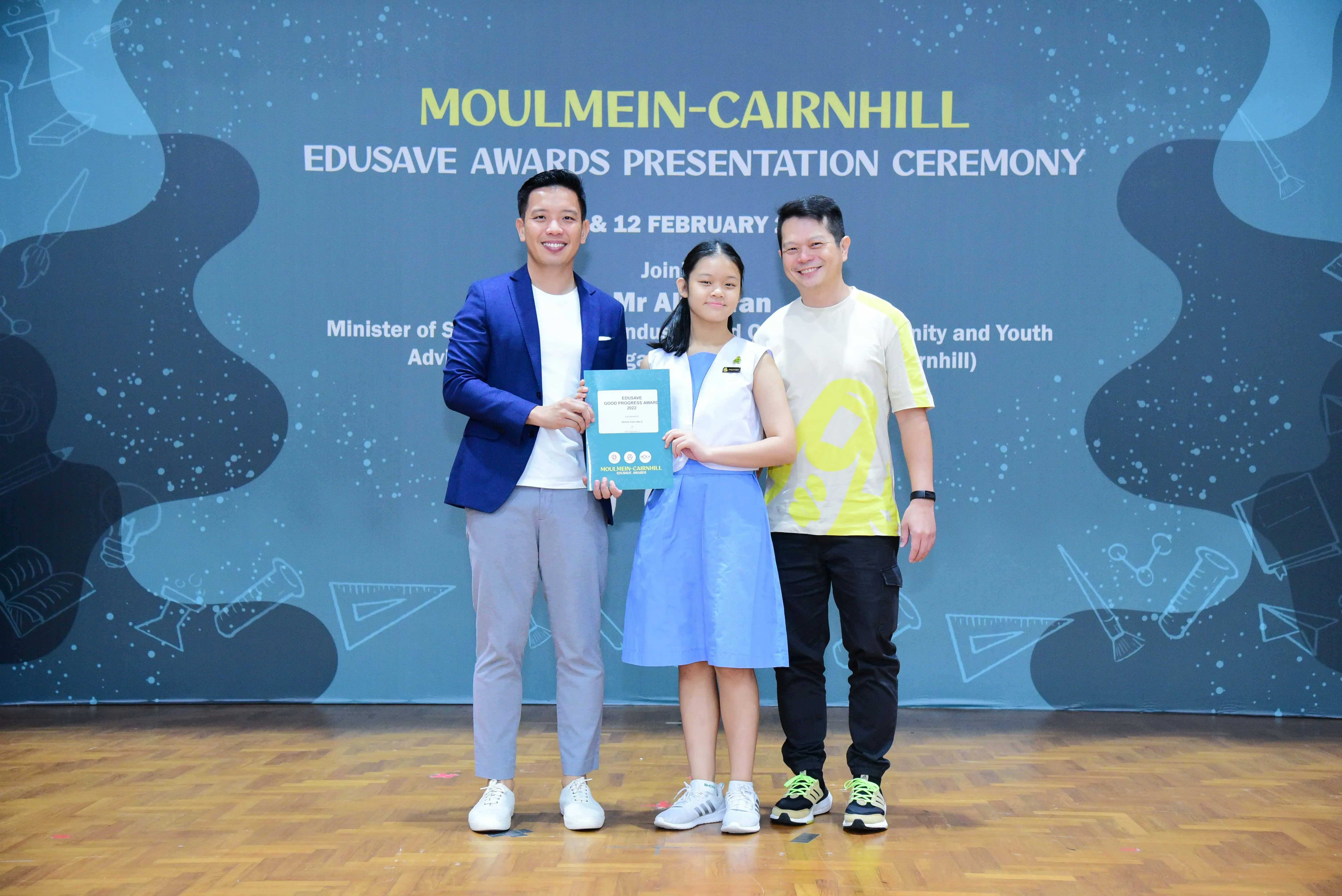 A girl holds an award certificate flanked by two smiling men at the Moulmein-Cairnhill Edusave Awards Presentation Ceremony.