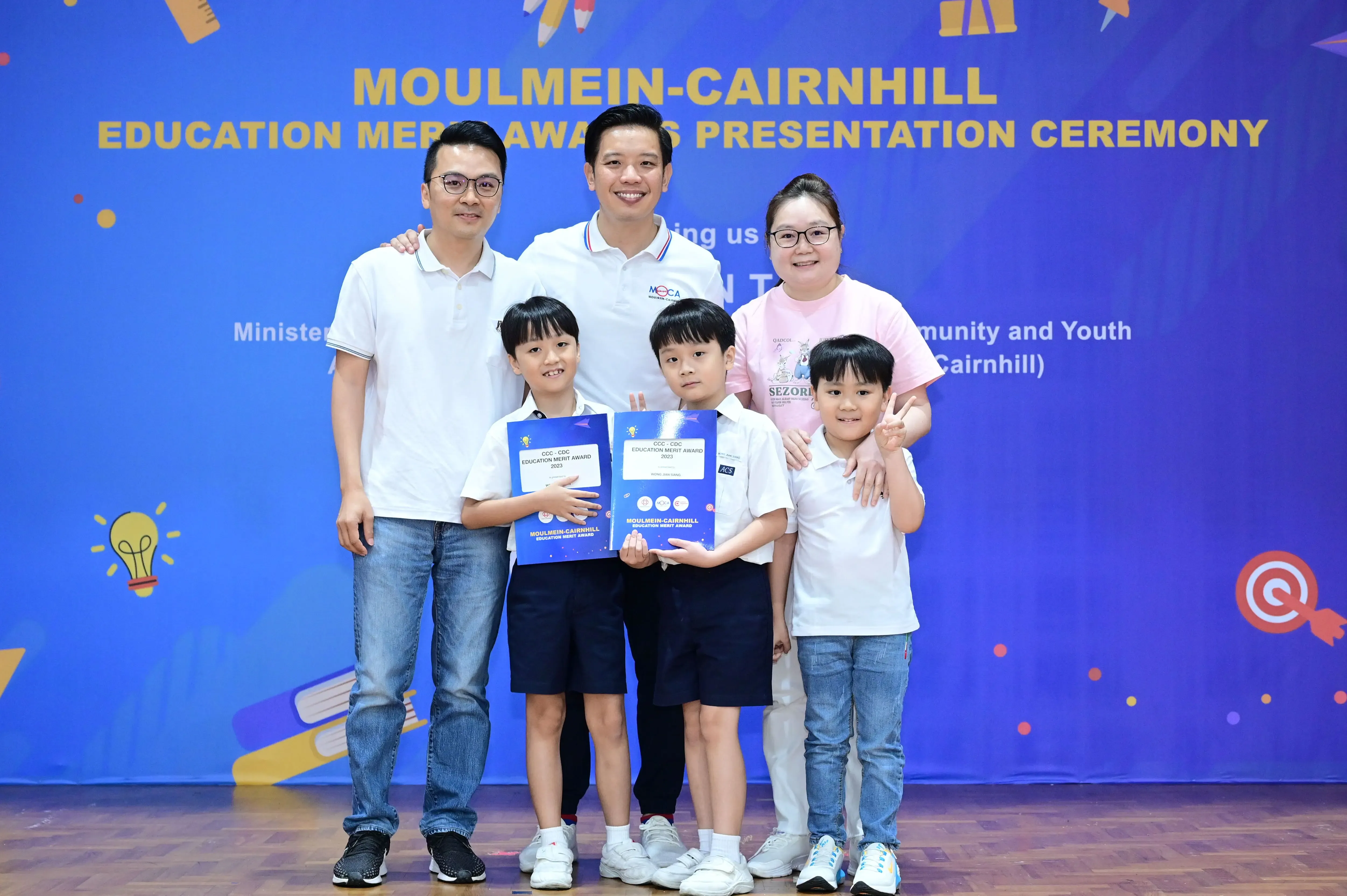 Three adults and three children pose at the Moulmein-Cairnhill Education Merit Awards Presentation Ceremony, two children holding award folders.