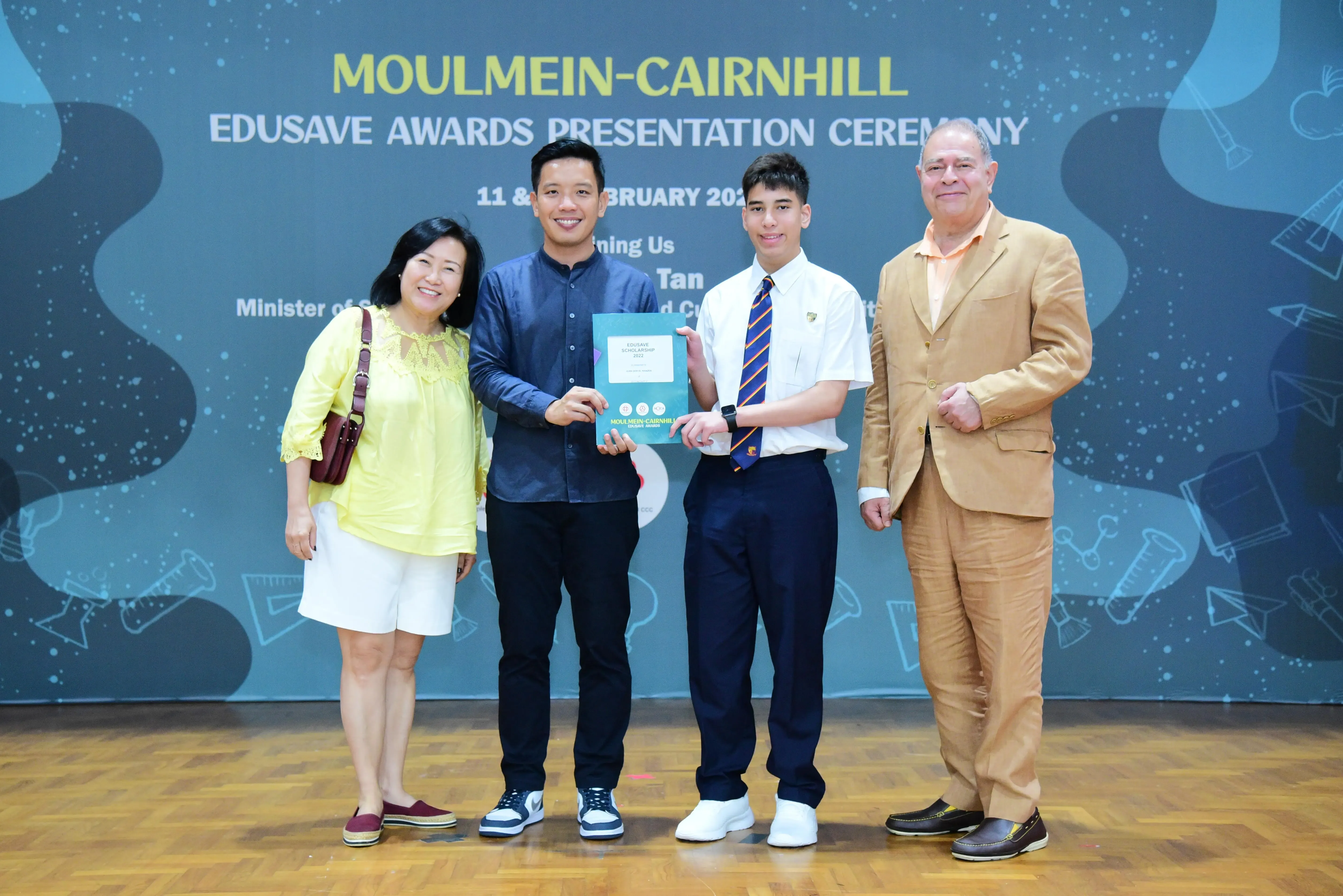 Four people stand smiling at the Moulmein-Cairnhill Edusave Awards Presentation Ceremony, with two holding a teal certificate folder.
